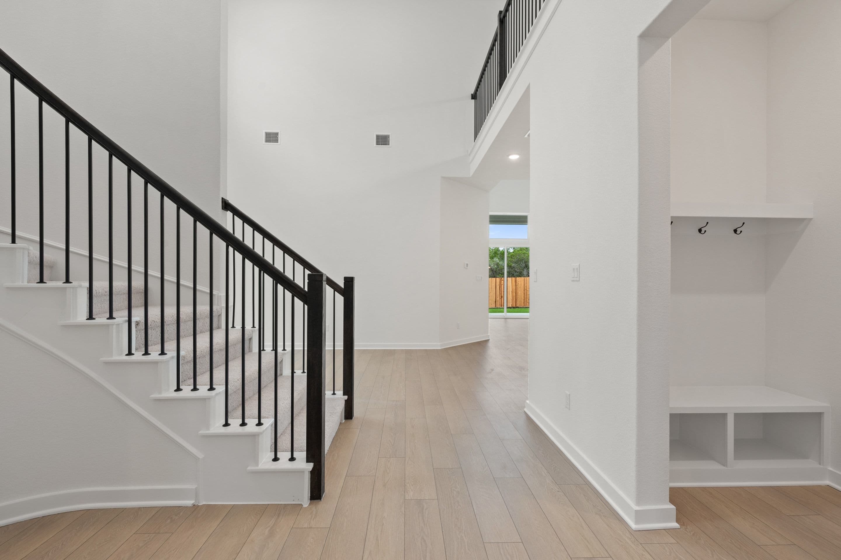 A modern staircase with black railings and a built-in bench with cubbies are in the foreground, while a bright hallway leading to a backyard with a wooden fence is in the background.