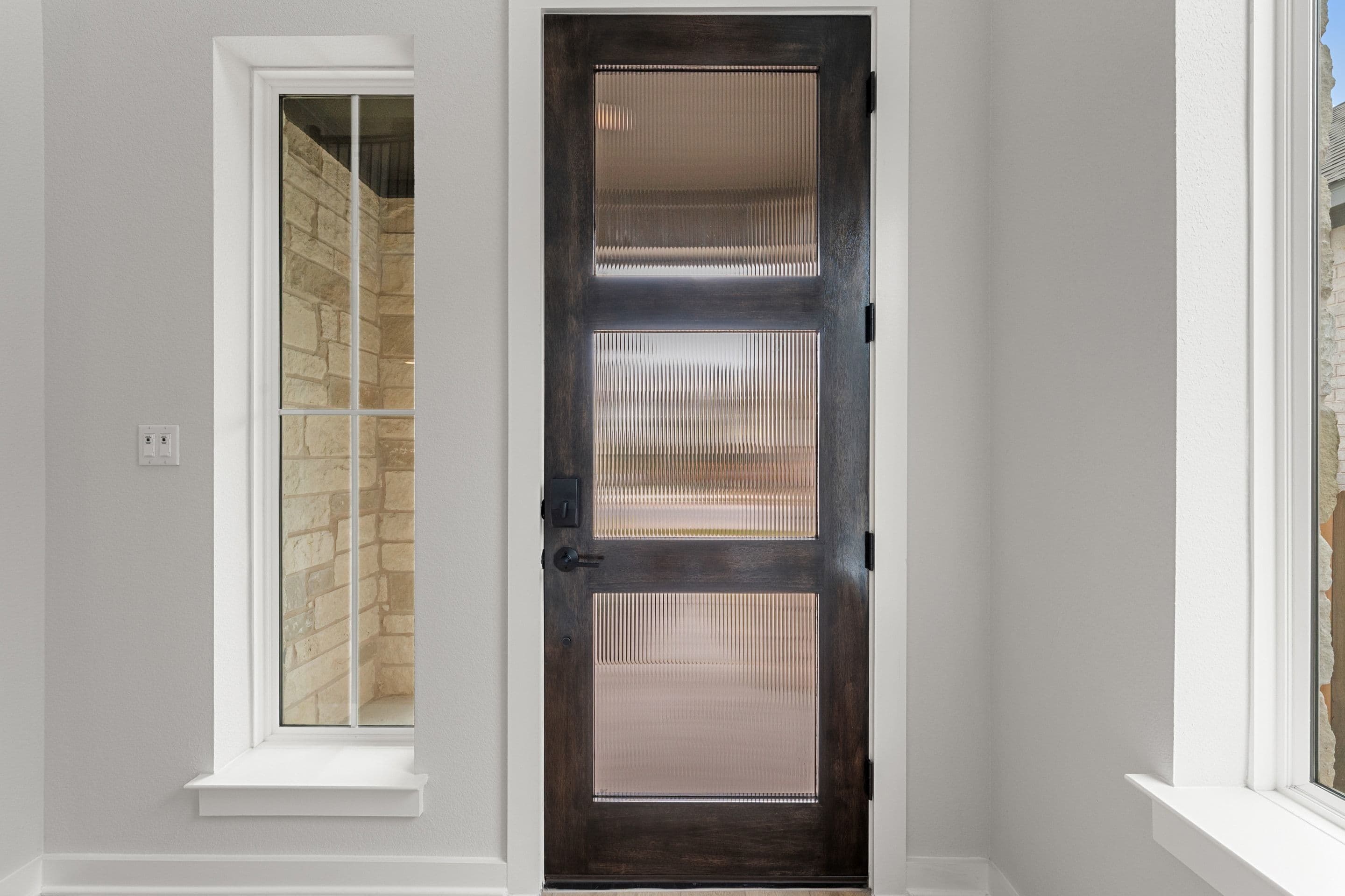 A dark wooden door with three glass panels stands in the foreground, while a window revealing a stone-tiled shower is visible to the left.