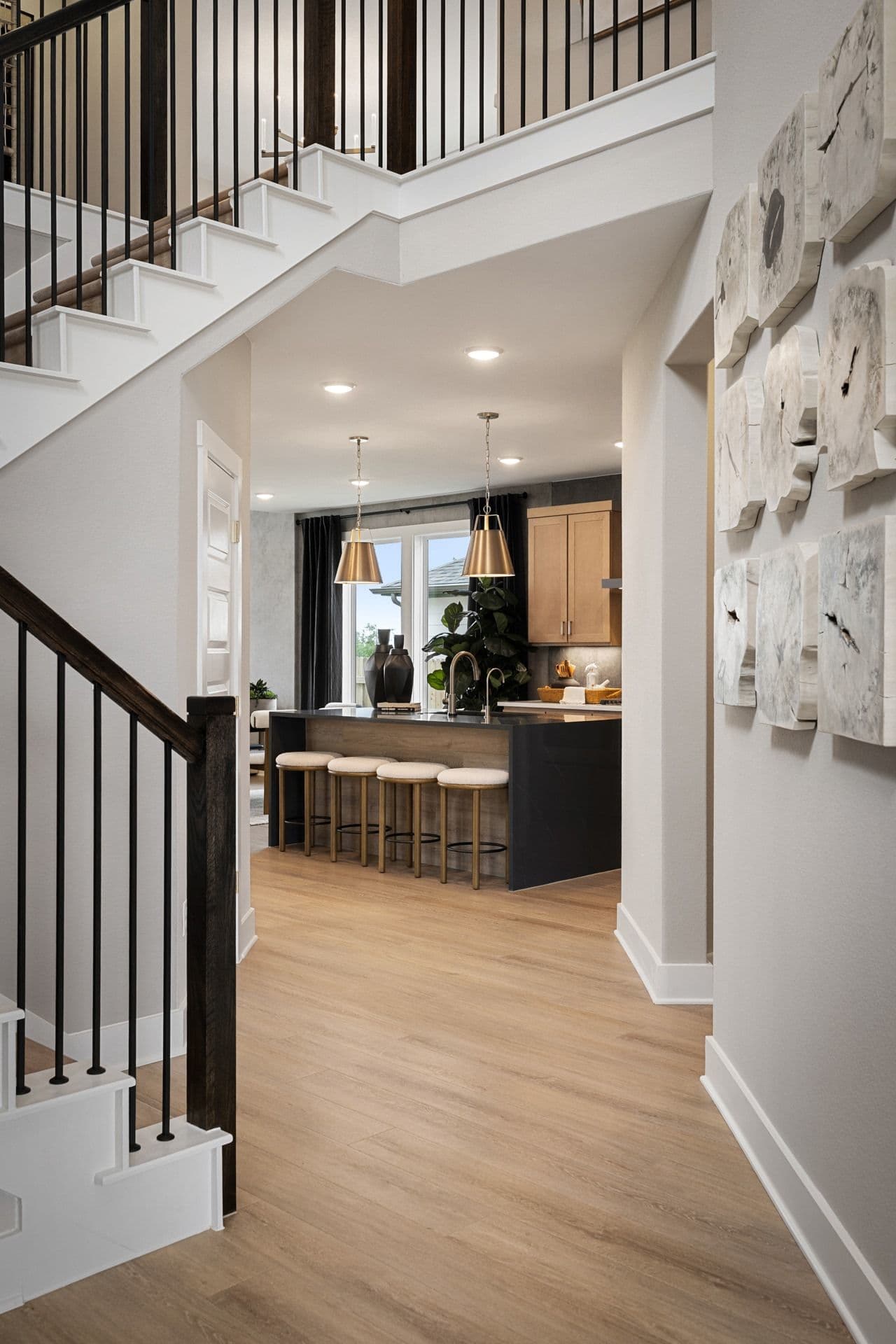 A modern staircase with dark wood railings leads to a bright kitchen with a large island and pendant lights, all set against a neutral-toned hallway.