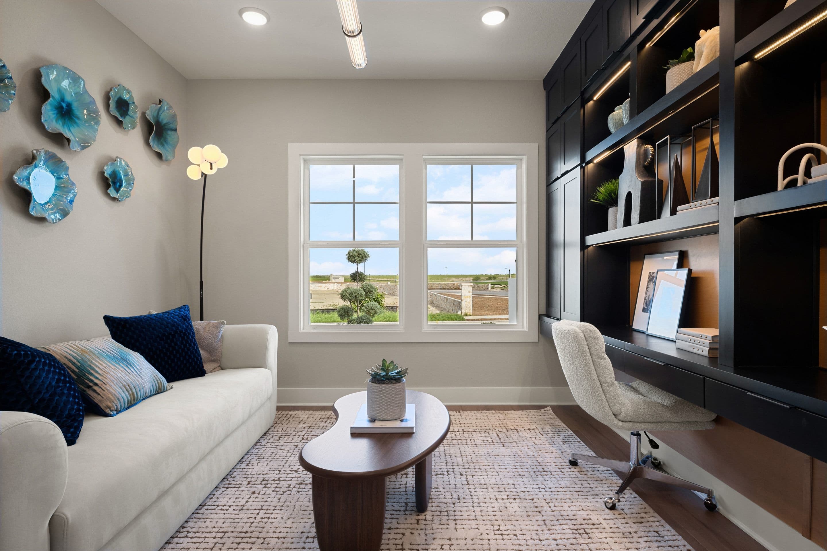 A modern office space features a white sofa and coffee table in the foreground, with a large window and built-in bookshelves in the background.