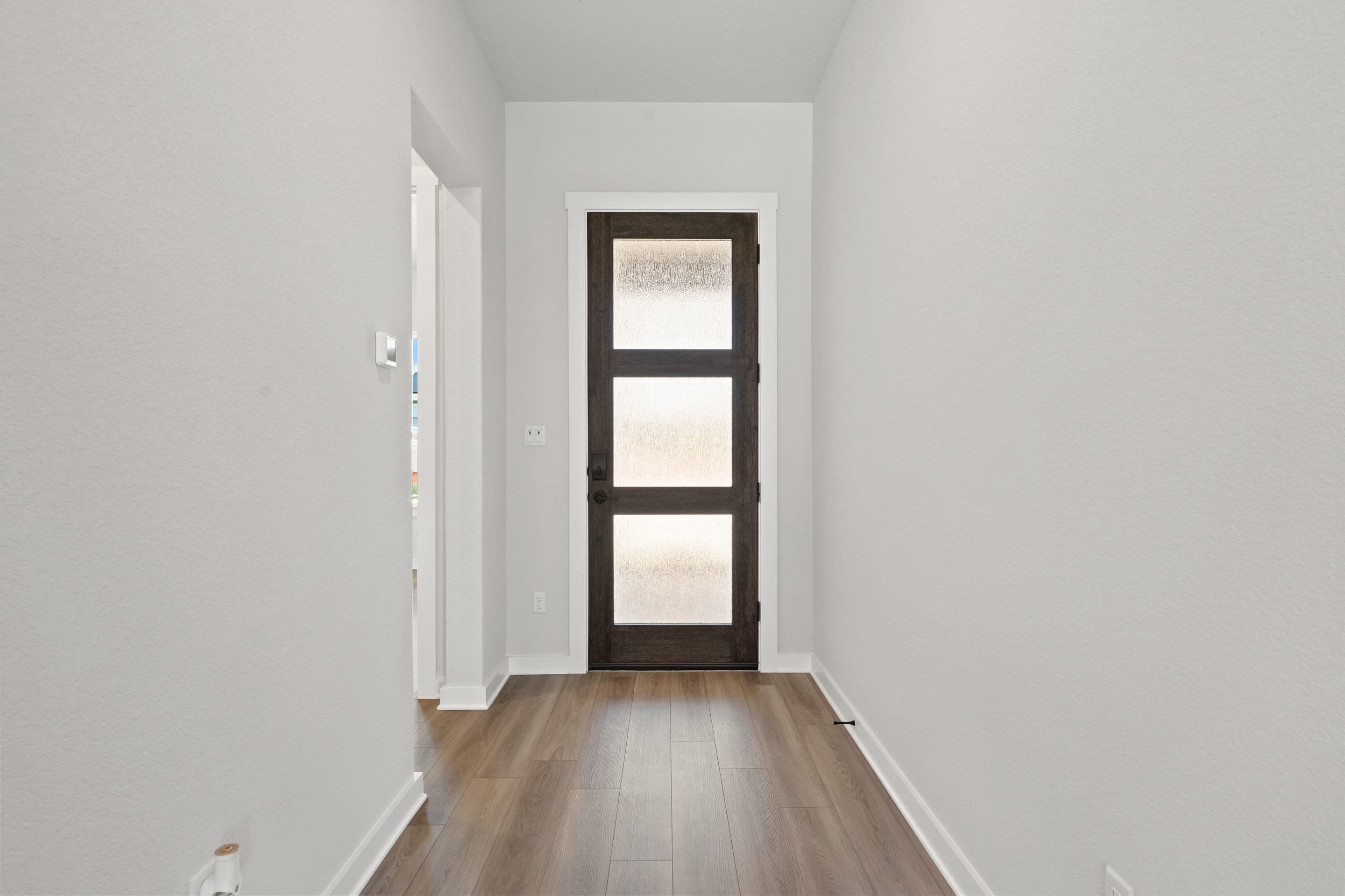 A narrow hallway with light gray walls and wood-look flooring leads to a dark brown door with three frosted glass panels.