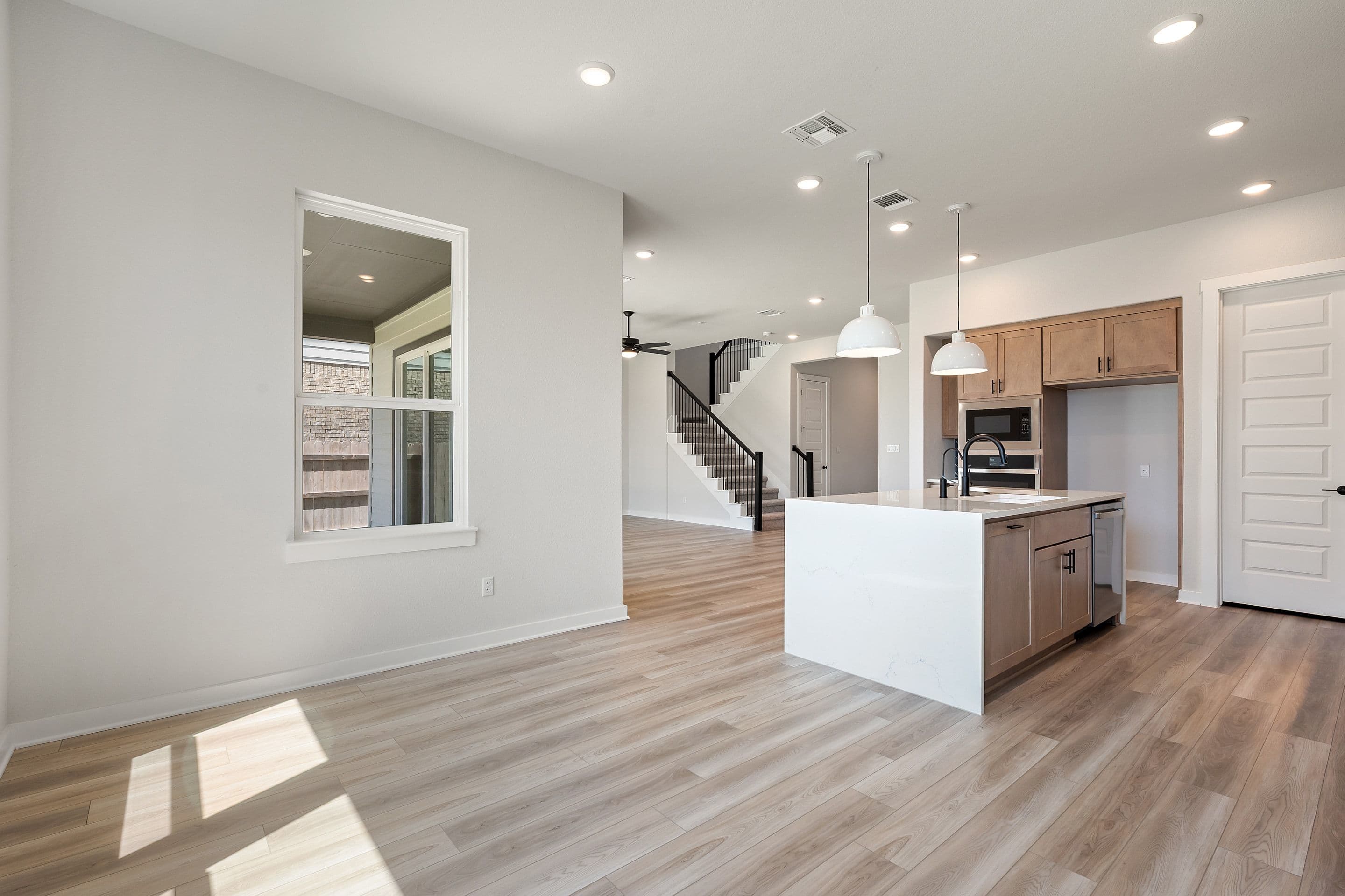 Sunlight streams across the light wood floor in the foreground, illuminating a modern kitchen island and cabinetry, while a staircase leads up to the background.