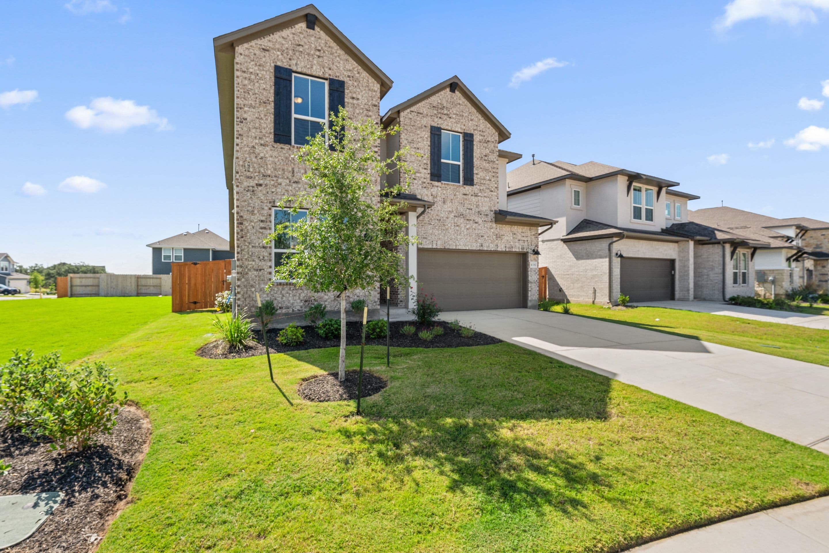 A modern brick house with a manicured lawn in the foreground sits in a suburban neighborhood with other similar houses in the background.