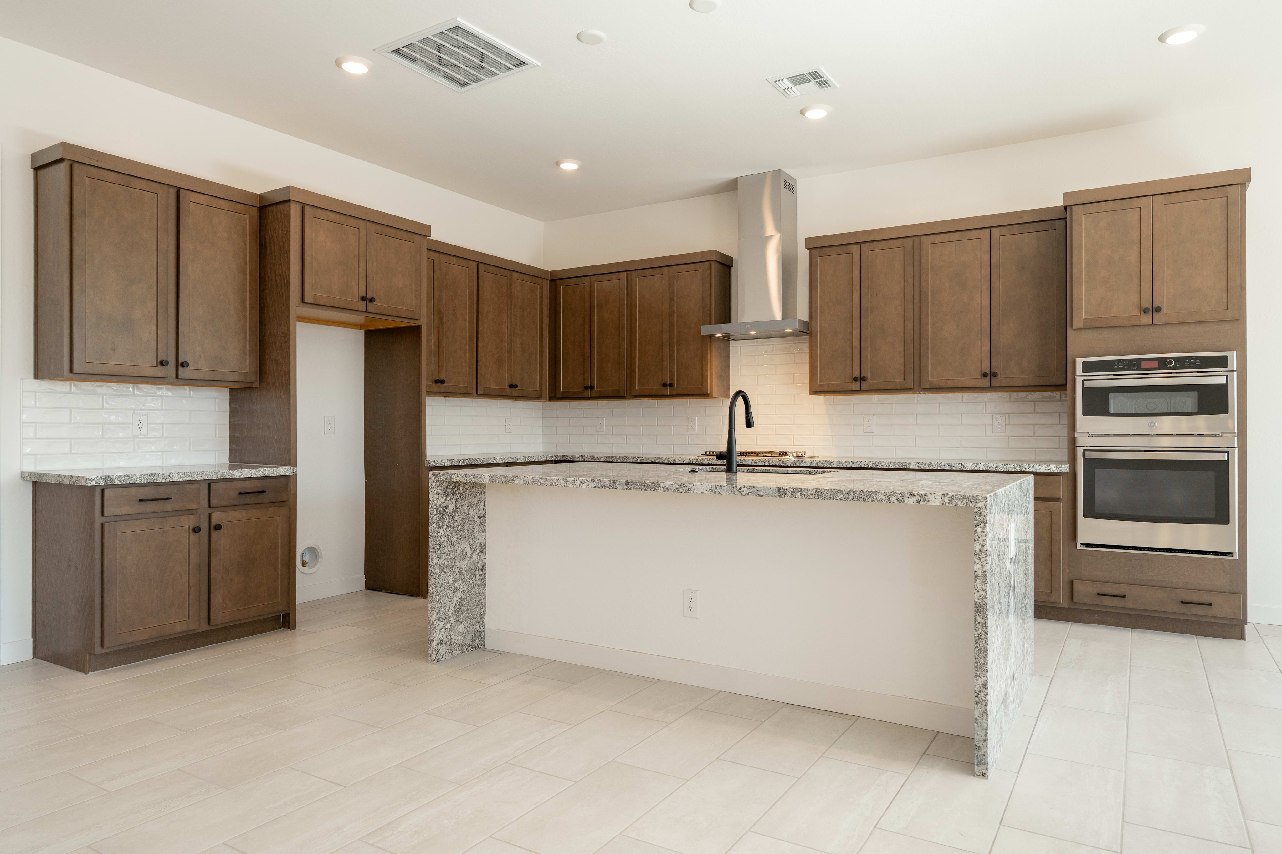 A modern kitchen with a granite island in the foreground and dark wood cabinets and stainless steel appliances in the background.