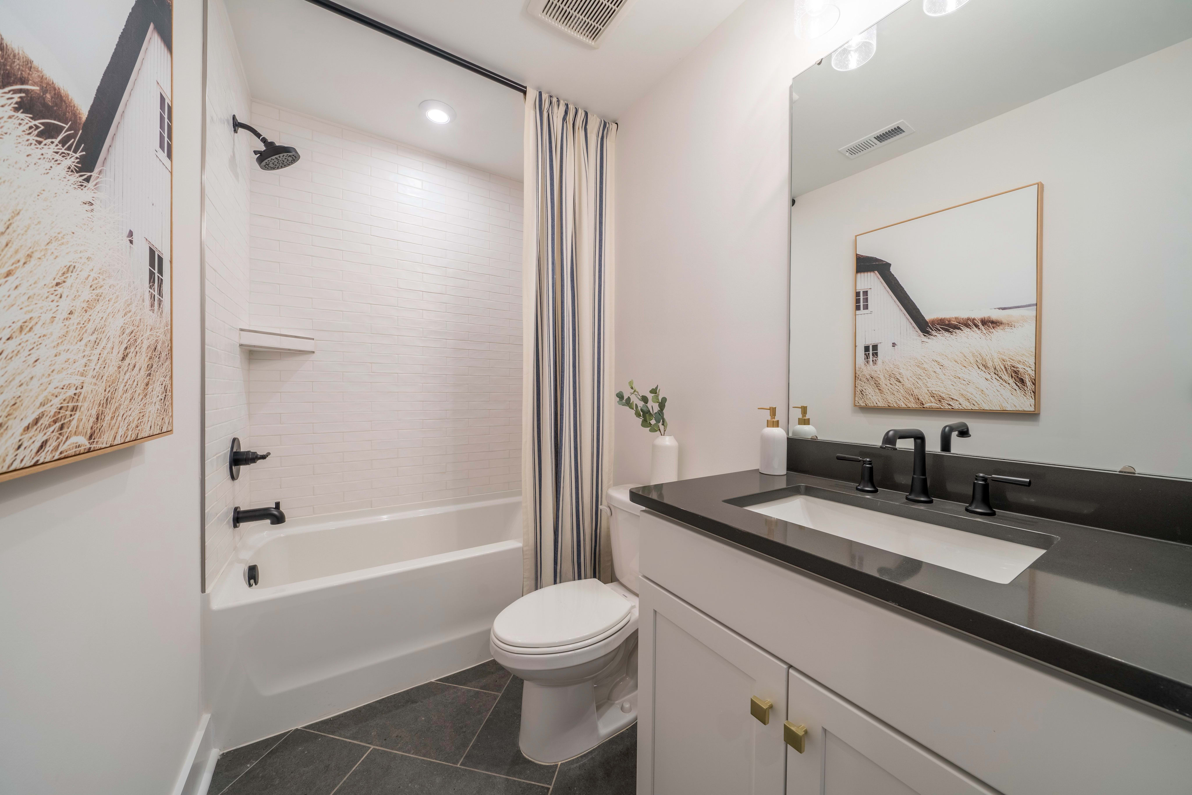 A modern bathroom features a white bathtub and toilet in the foreground, with a vanity and mirror against a wall adorned with artwork in the background.