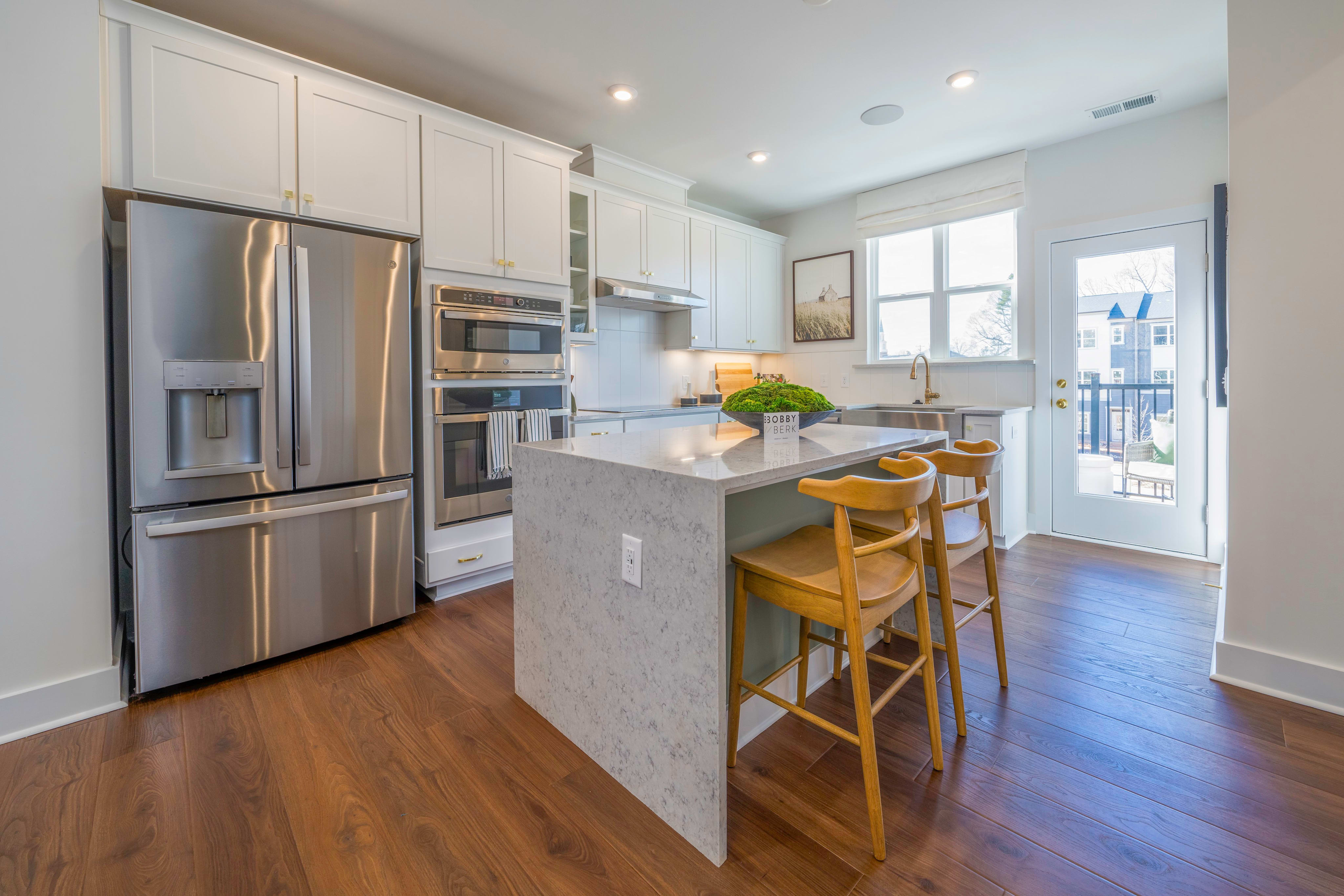 A modern kitchen with a stainless steel refrigerator and island with bar stools in the foreground, and a window and door leading to an outdoor space in the background.