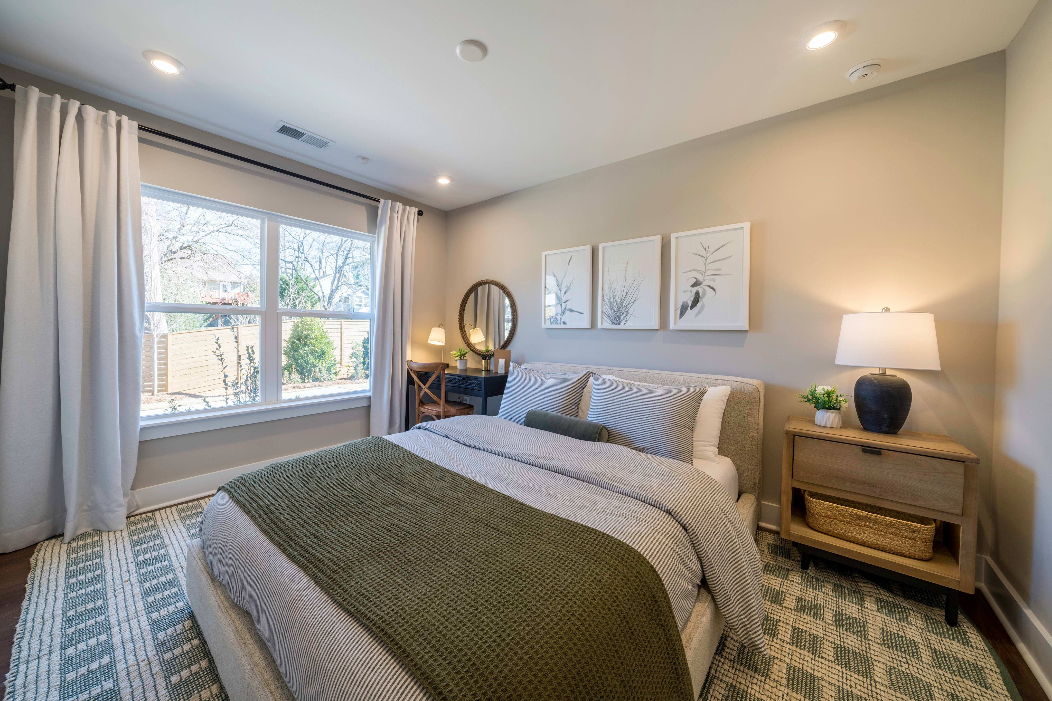 A neatly made bed with a green throw blanket is in the foreground, while a window with white curtains and a desk with a mirror are in the background.