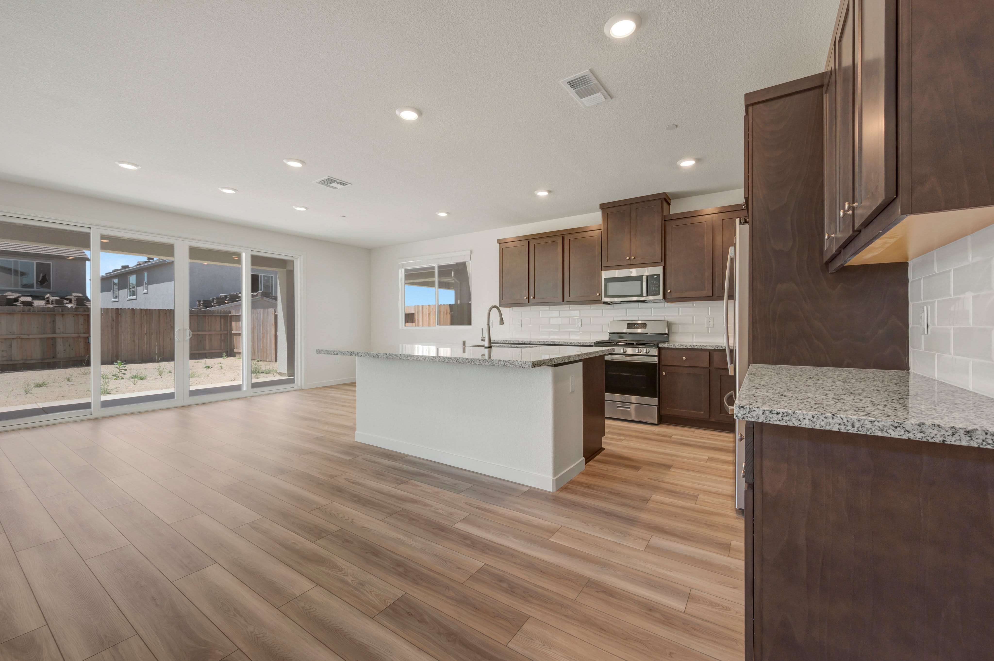 A modern kitchen with dark wood cabinets and a large island in the foreground opens to a bright living area with sliding glass doors leading to a backyard in the background.