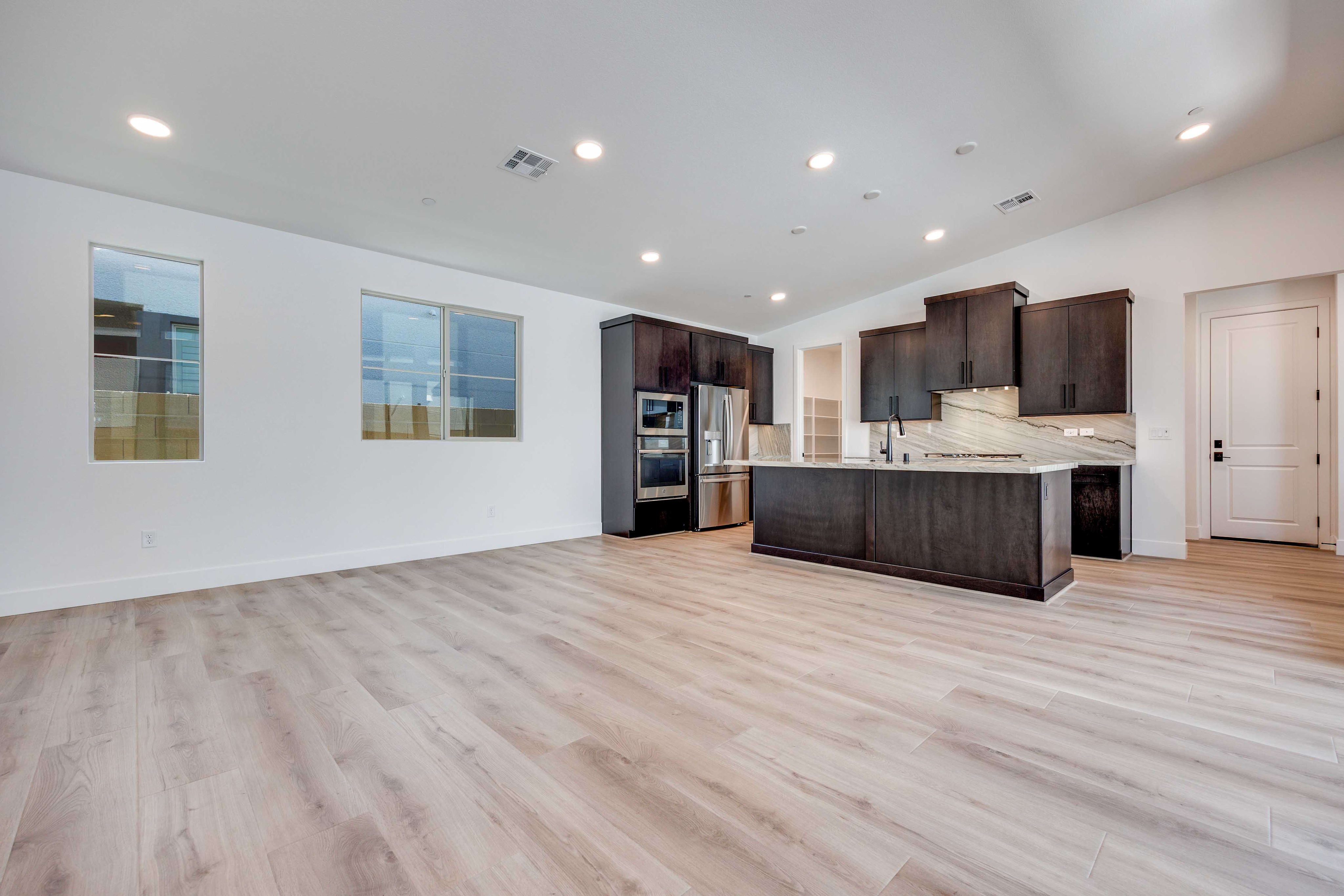 A spacious, modern living area with light wood flooring in the foreground opens to a dark-cabinet kitchen with stainless steel appliances and a marble backsplash in the background.