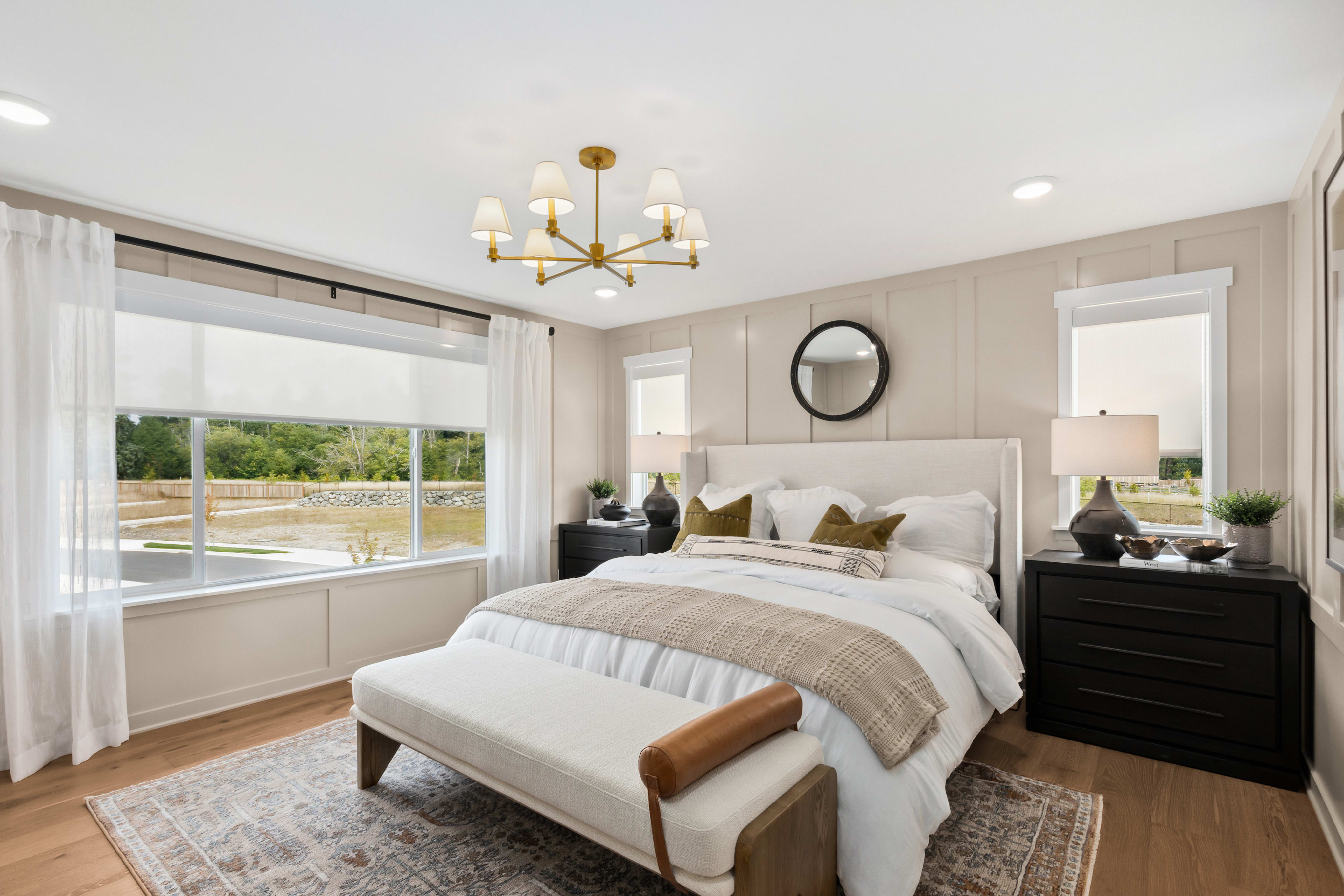 A well-lit bedroom with a large bed in the foreground and a window overlooking a green landscape in the background.