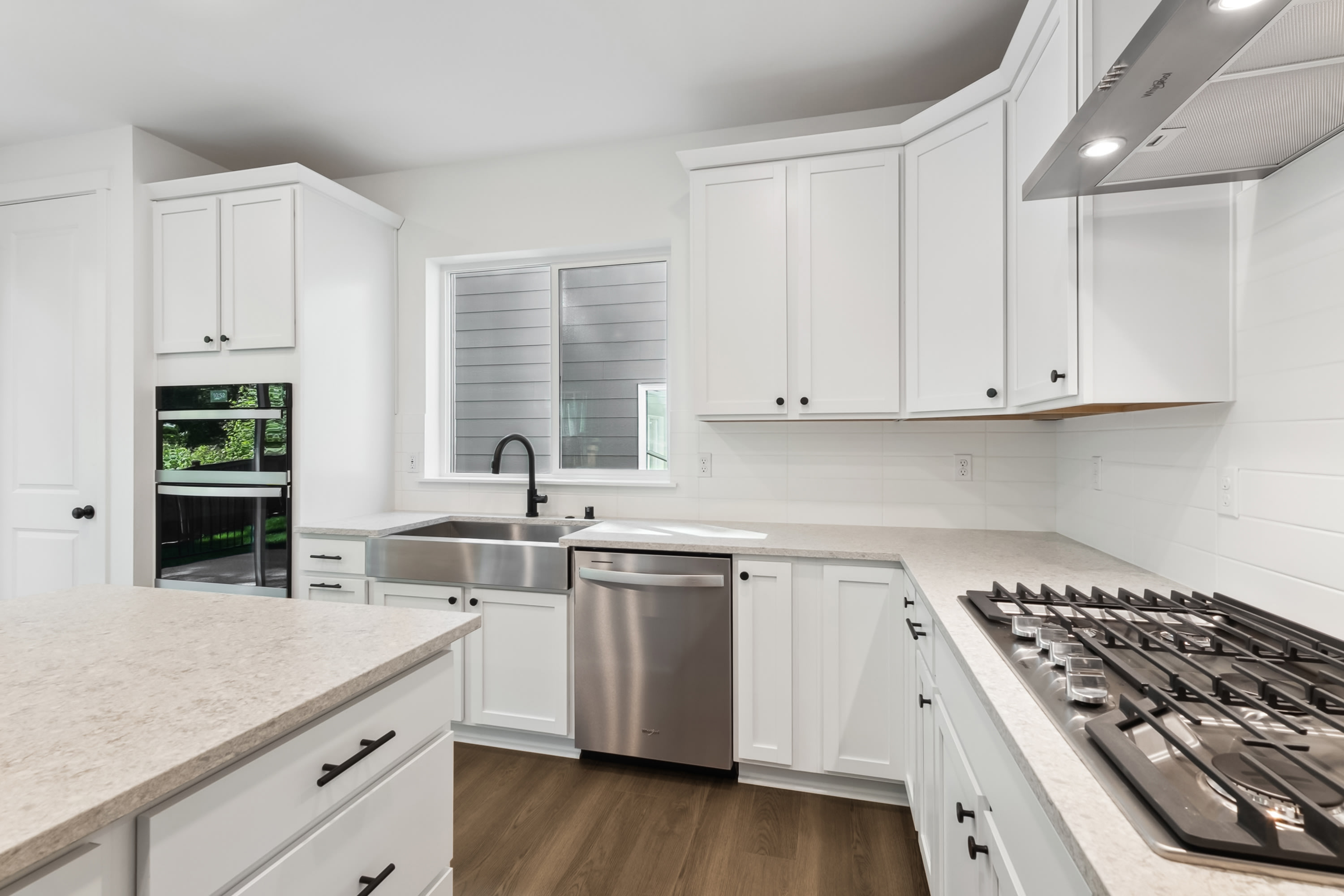 A modern white kitchen with a granite countertop island in the foreground and stainless steel appliances and white cabinetry in the background.