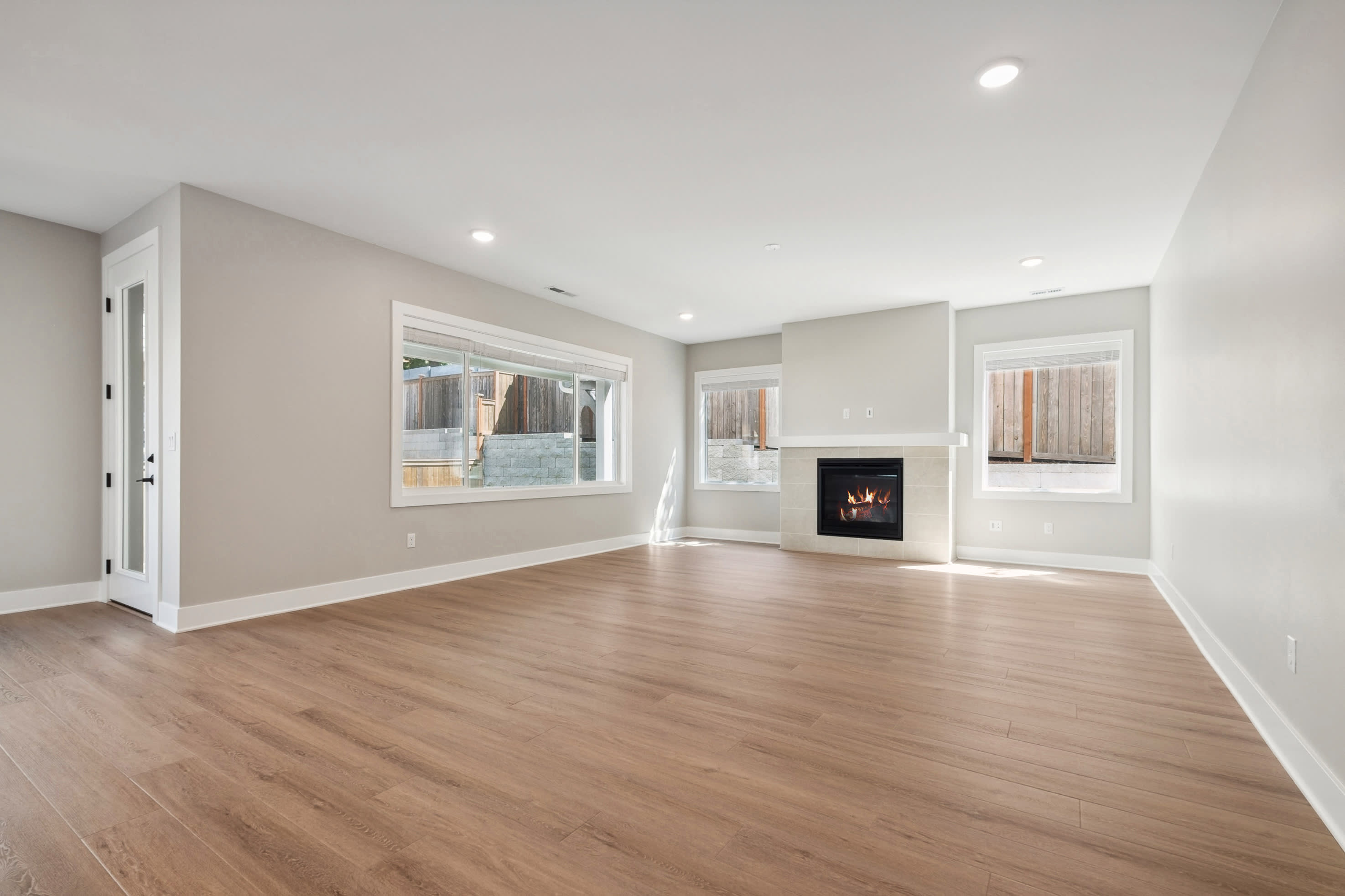A spacious, modern living room with light wood floors in the foreground and a fireplace with windows looking out to a fenced yard in the background.