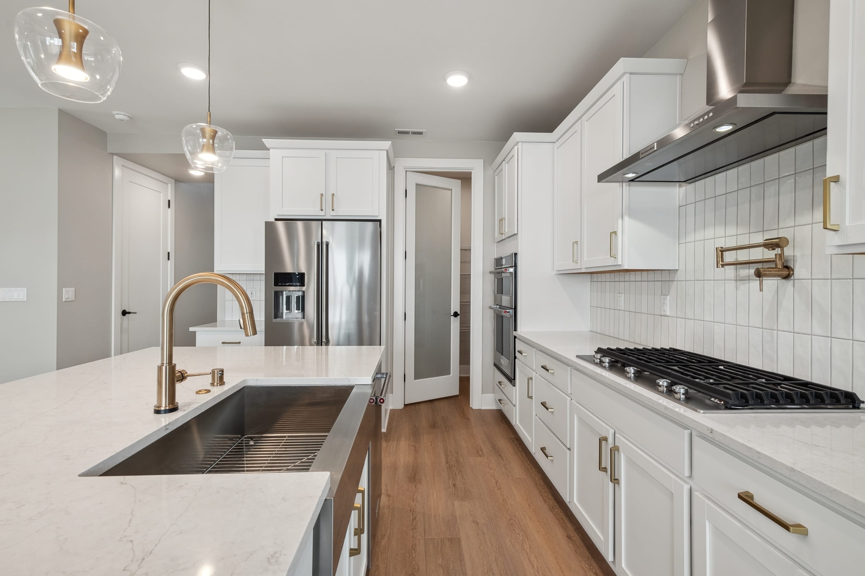 A modern white kitchen with a large island and sink in the foreground, and stainless steel appliances and a gas range in the background.