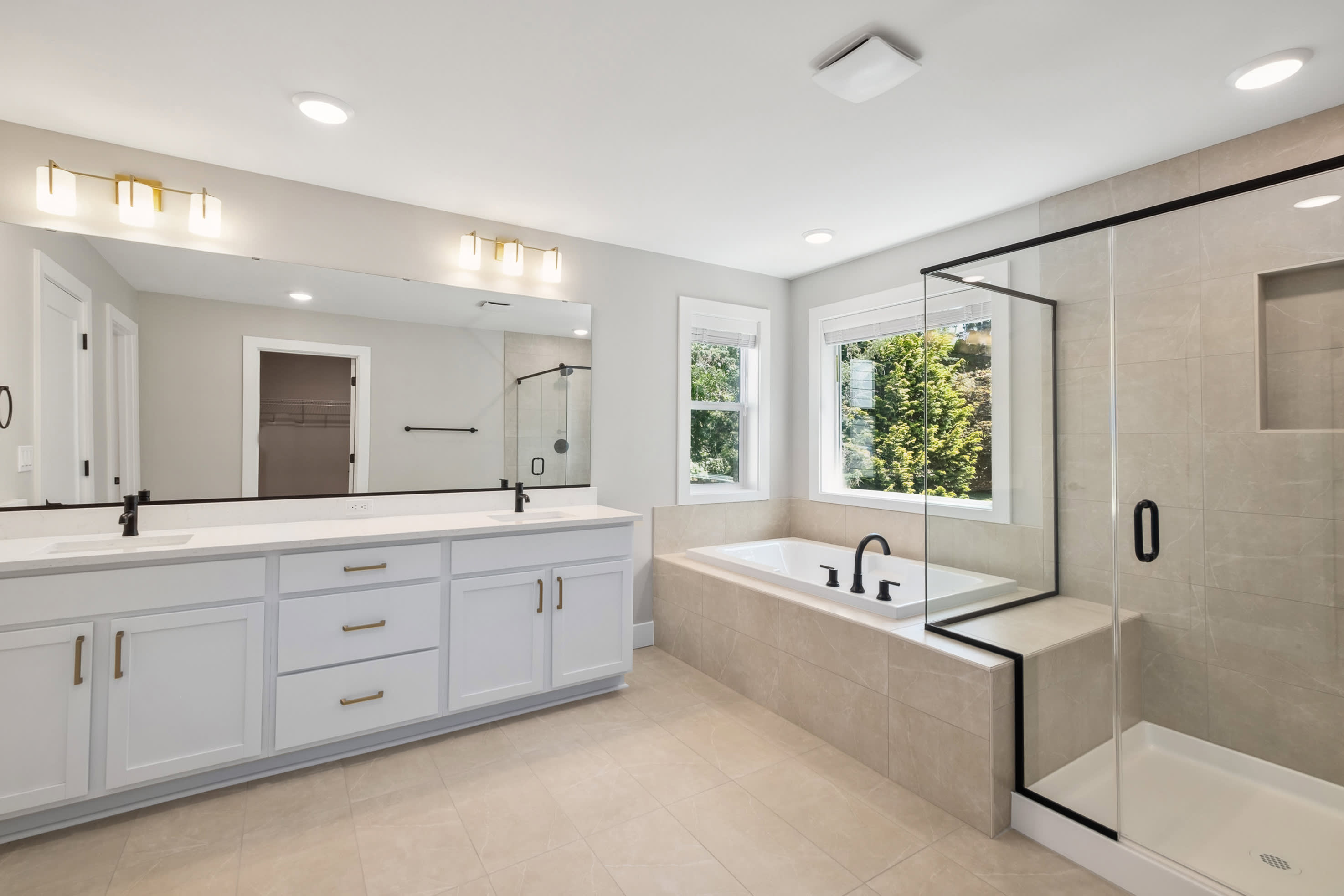 A modern bathroom features a double vanity with a large mirror in the foreground, and a freestanding bathtub and glass-enclosed shower in the background.
