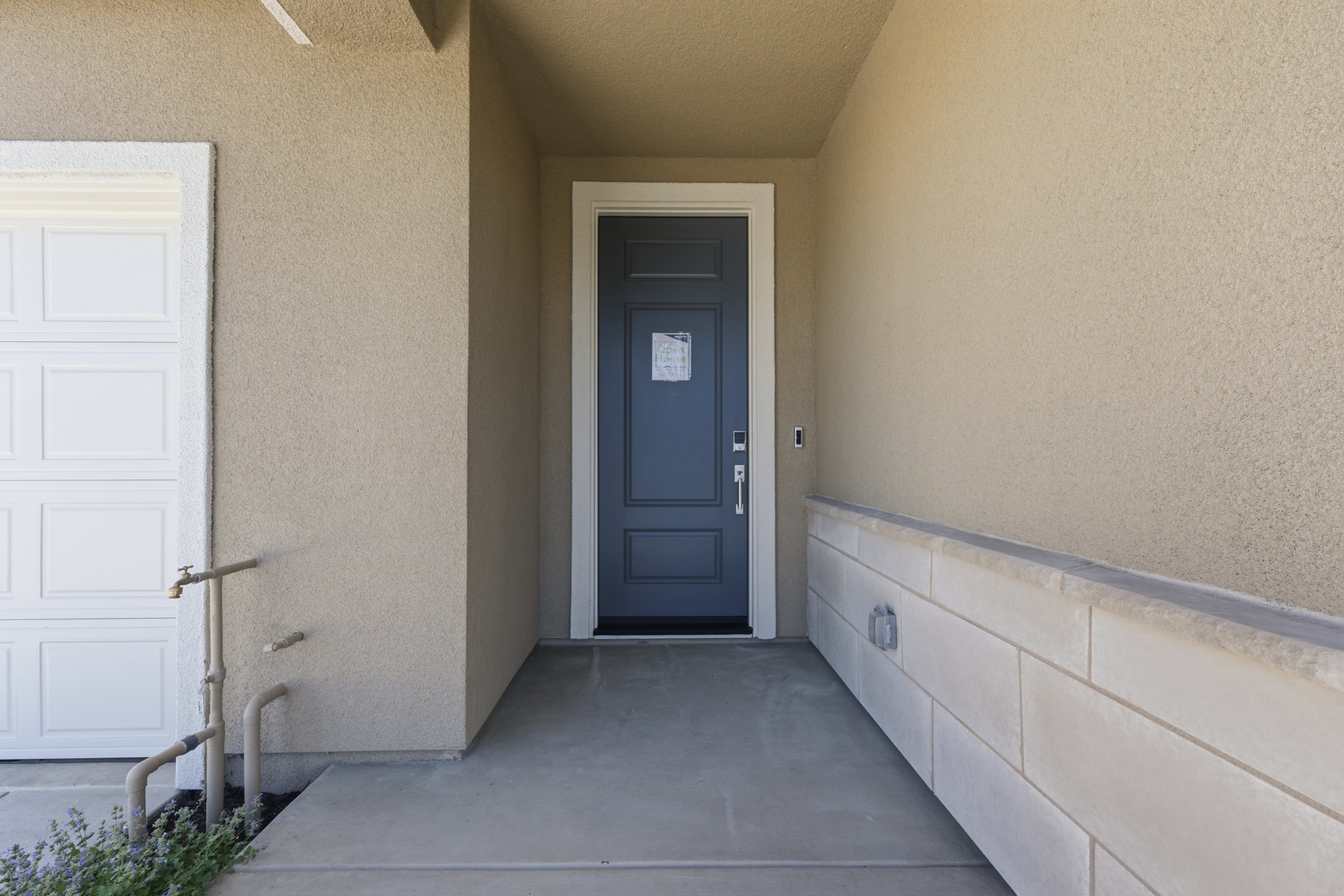 A gray front door with a white frame is centered in the background, while the foreground features a concrete walkway leading to it, bordered by a textured wall on the right and a white garage door on the left.