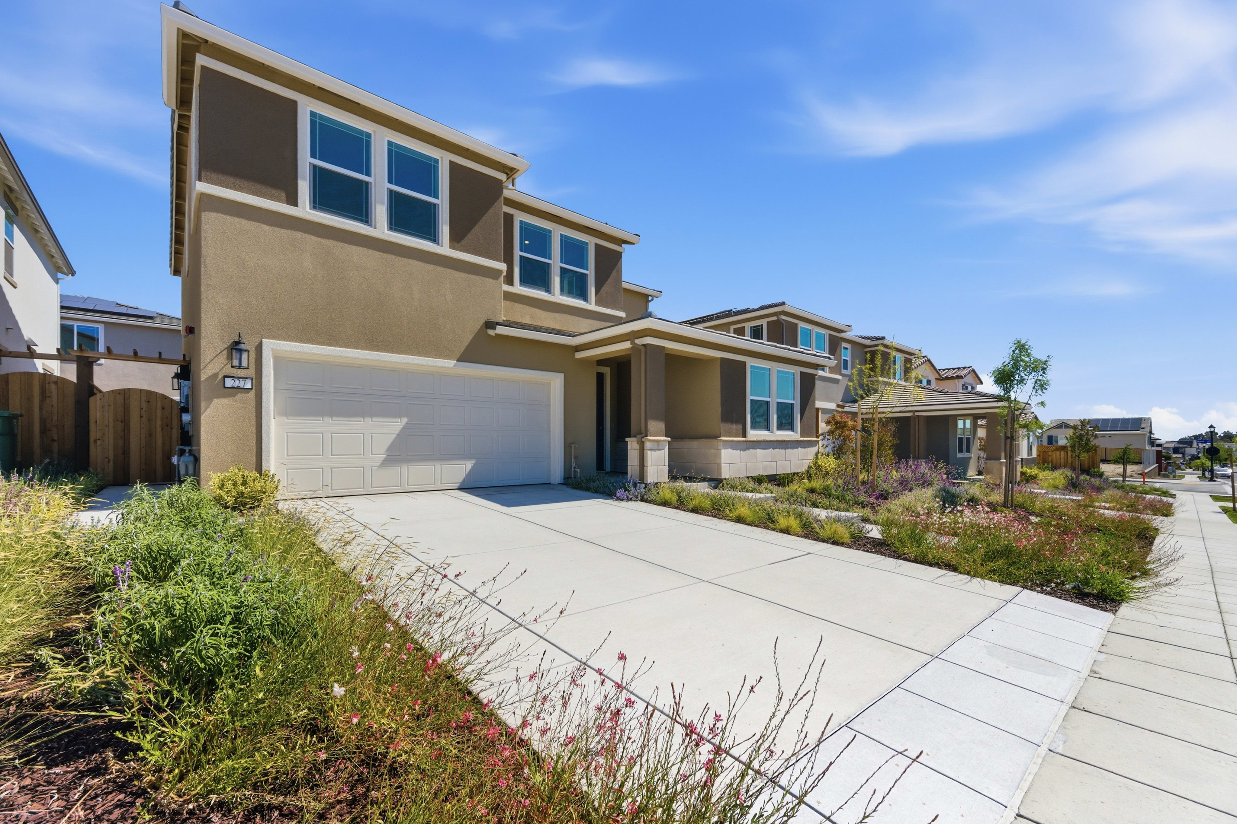 A modern two-story house with a beige exterior and a white garage door sits on a concrete driveway, bordered by a sidewalk and a landscaped yard with colorful flowers, all under a bright blue sky with scattered clouds.