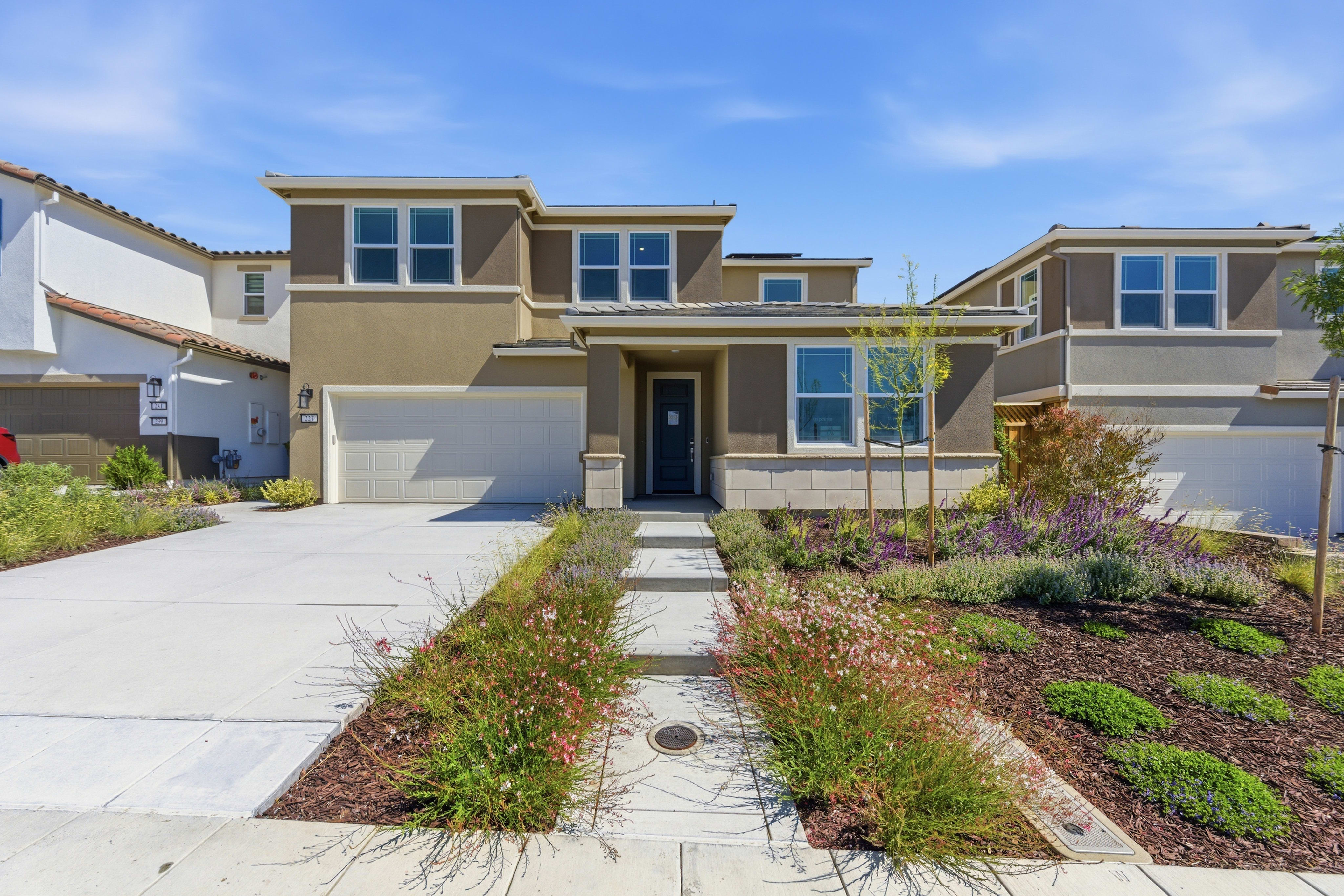 A modern two-story house with a well-maintained front yard featuring a pathway and colorful flowers is set against a clear blue sky.