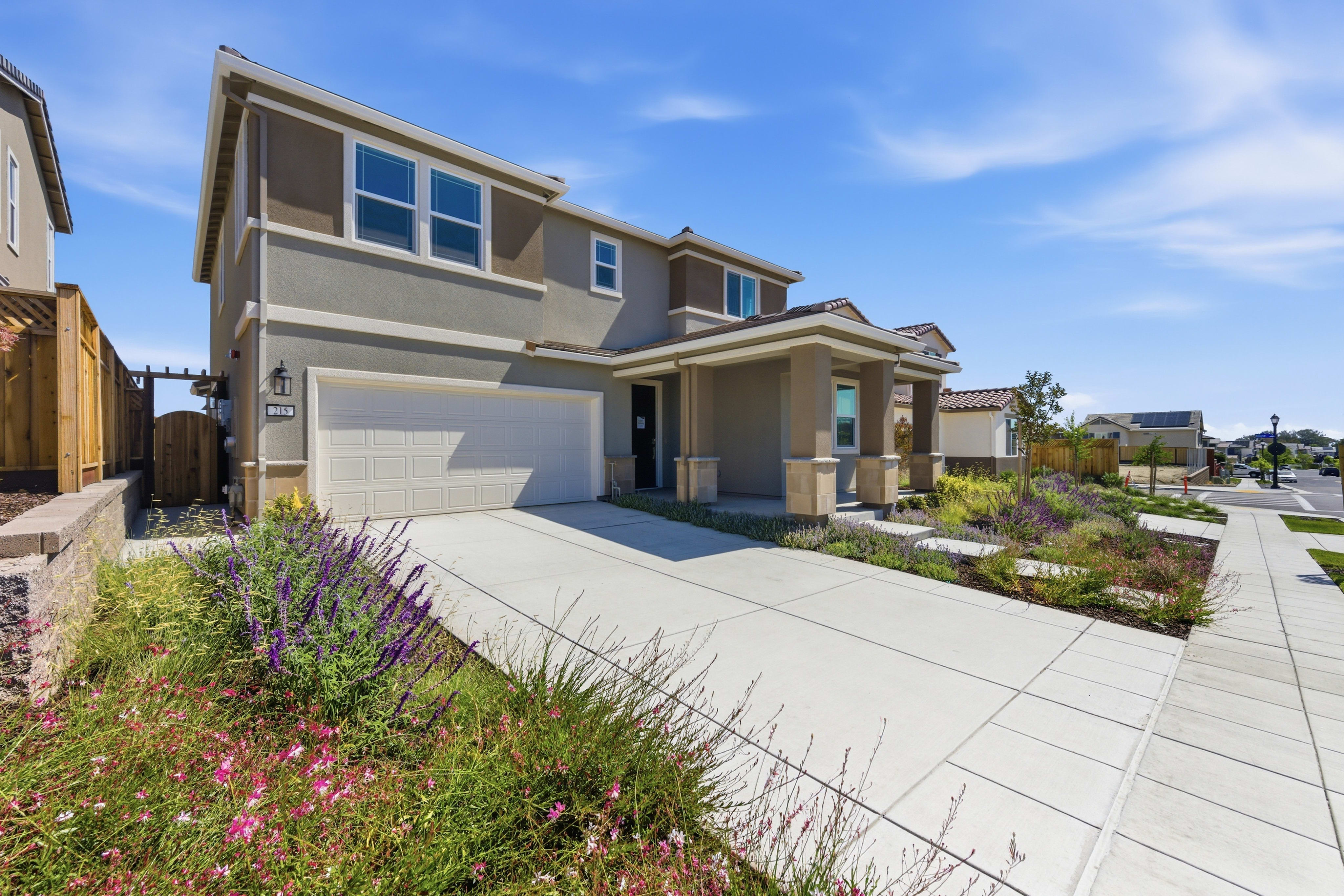A modern two-story house with a light brown exterior and a white garage door sits on a concrete driveway, bordered by a colorful array of wildflowers and manicured landscaping, with a clear blue sky and distant houses in the background.