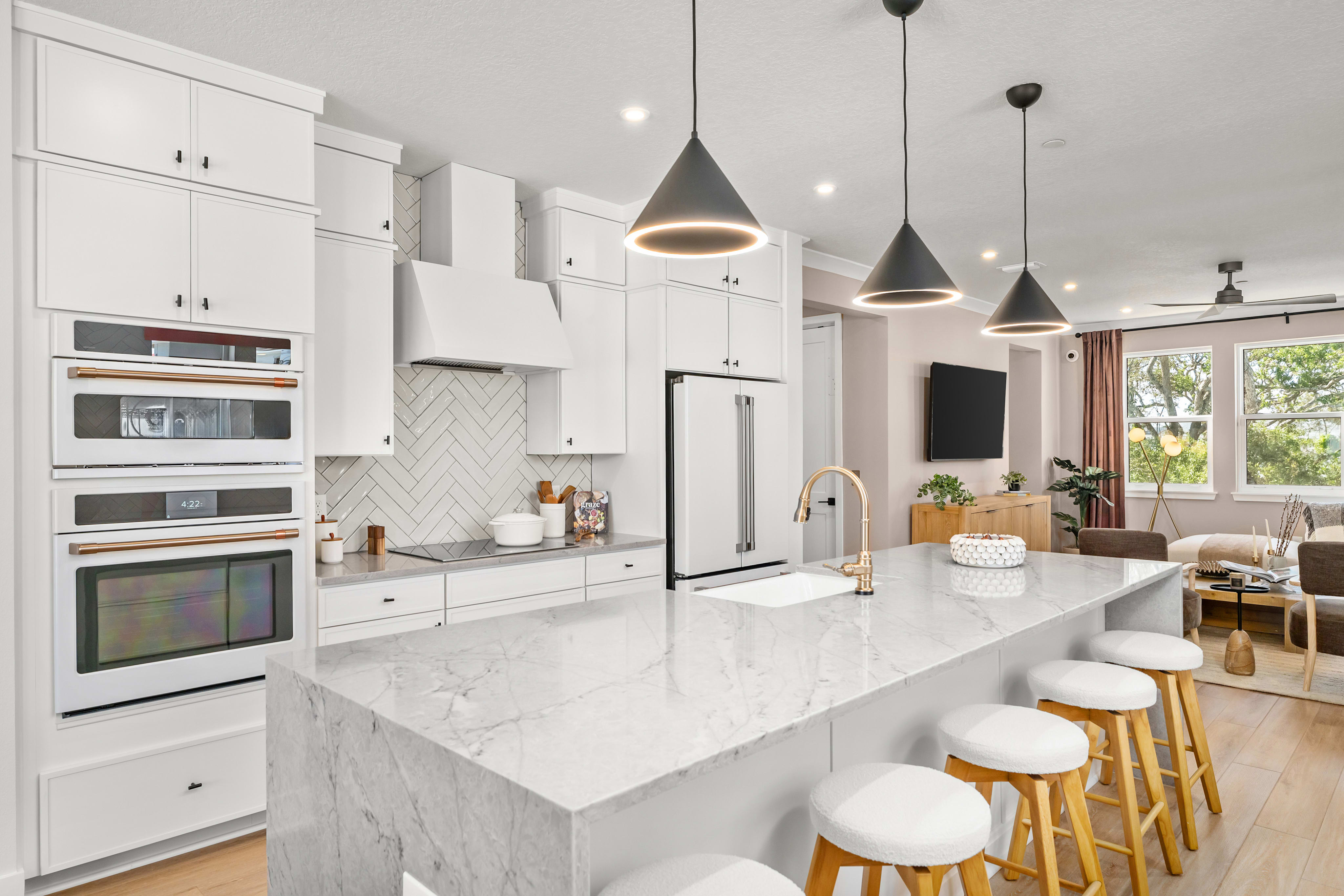 A modern white kitchen island with marble countertops and pendant lights is in the foreground, while a living area with a television and large windows is visible in the background.