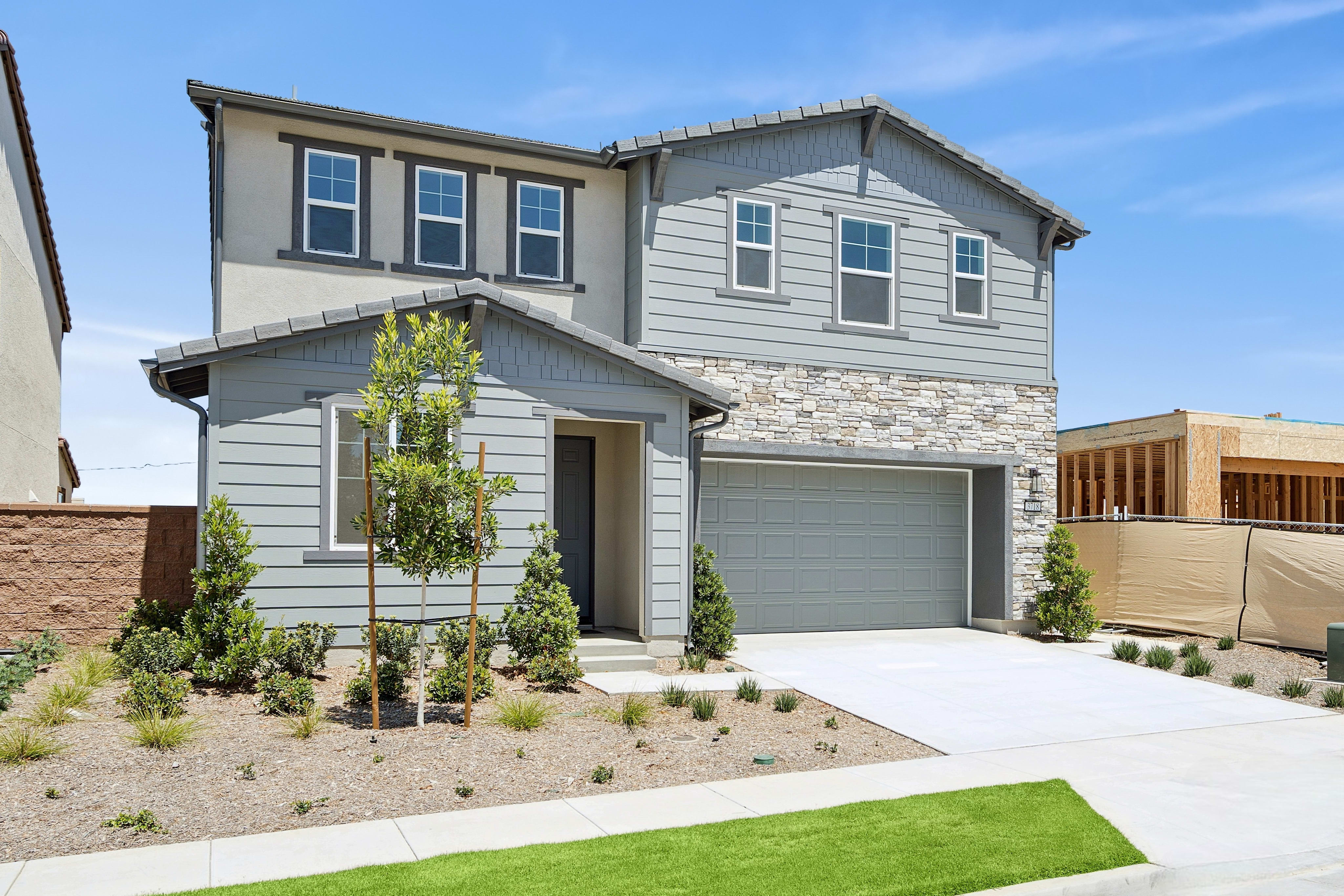 A modern two-story house with a stone and siding facade sits in the foreground, while a construction site with wooden framing and fencing is visible in the background under a clear blue sky.