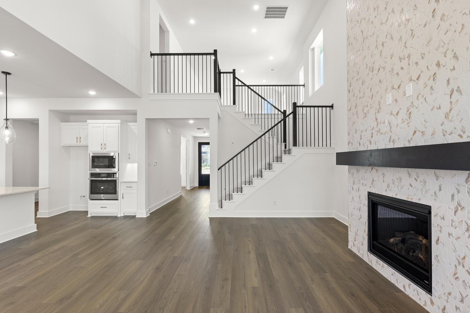 A modern living room with a fireplace and a staircase in the foreground, and a kitchen and hallway in the background.