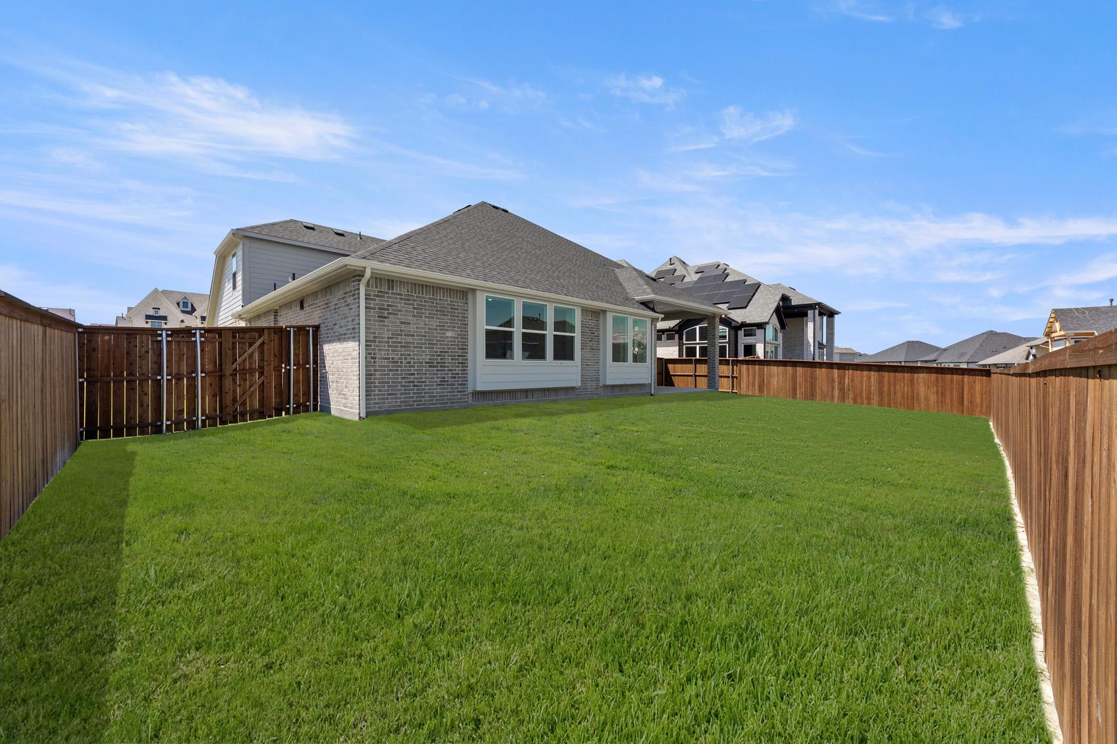 A freshly mowed green lawn fills the foreground, leading to a modern brick house with large windows and a covered patio in the background, all enclosed by a wooden fence under a bright blue sky.