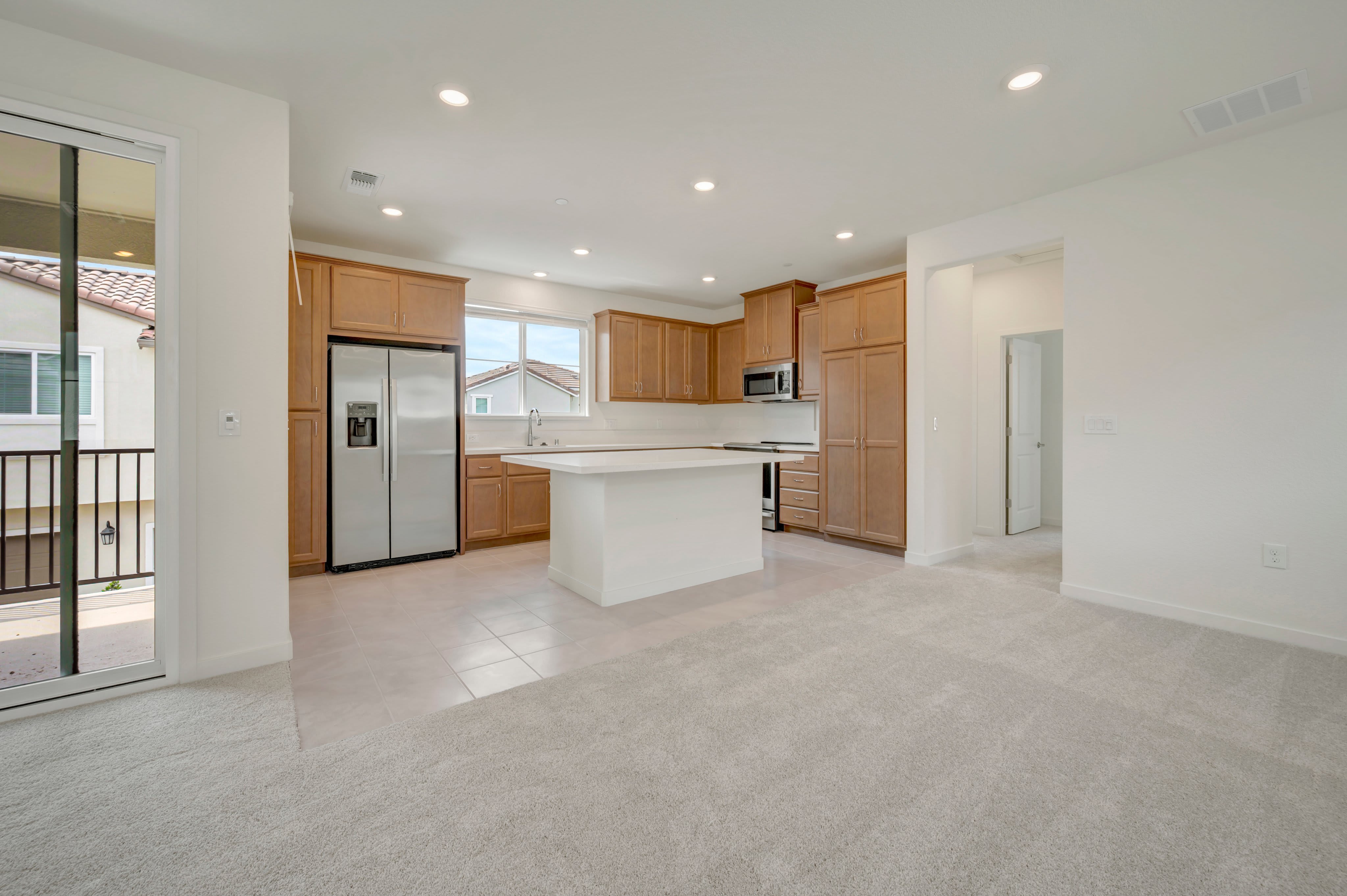 A spacious, modern kitchen with a large island and stainless steel appliances is visible in the background, while the foreground is a plush, neutral-toned carpeted living area with a sliding glass door leading to a balcony.