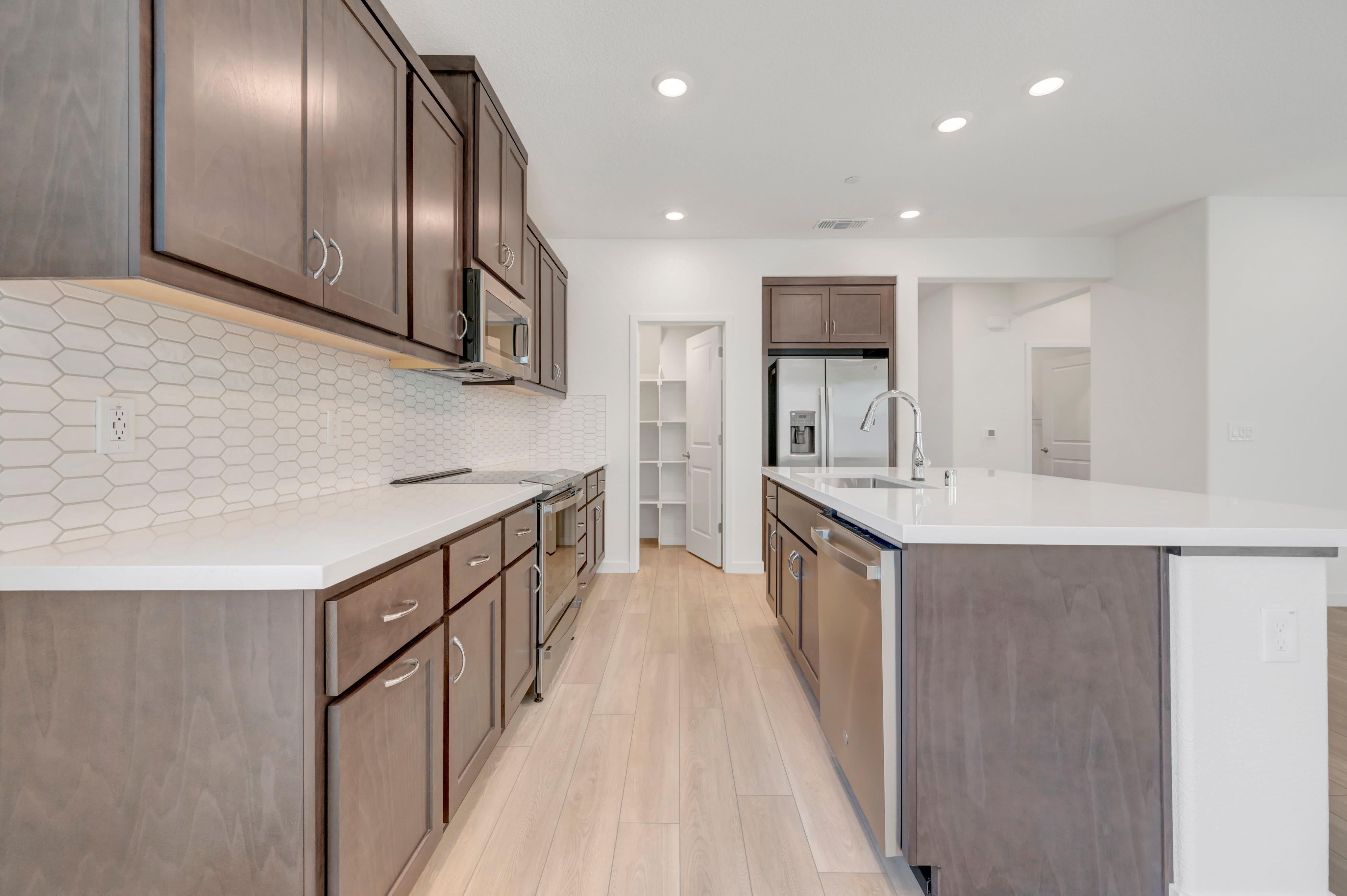 A modern kitchen with dark wood cabinets and a white tiled backsplash in the foreground, and a pantry and stainless steel refrigerator in the background.