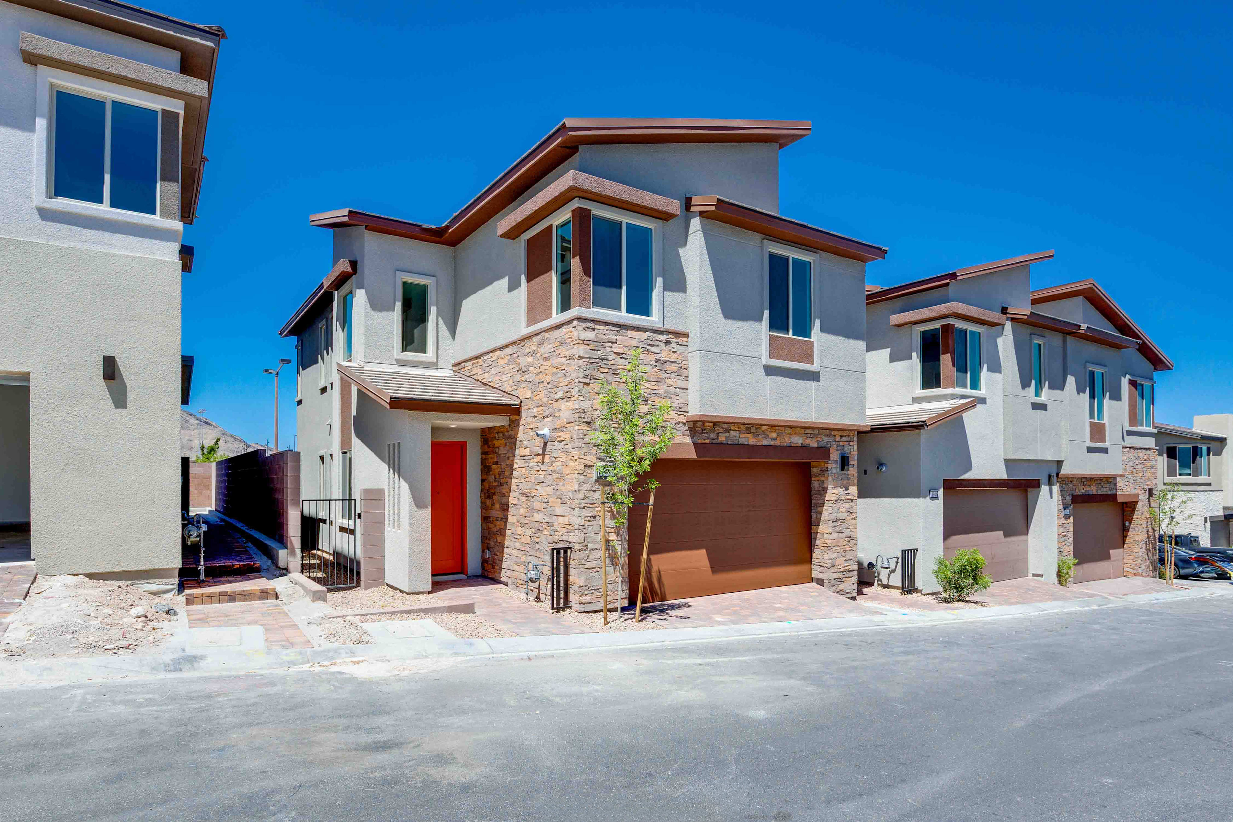 A row of modern townhouses with stone accents and orange doors stands under a clear blue sky, with a paved street in the foreground.