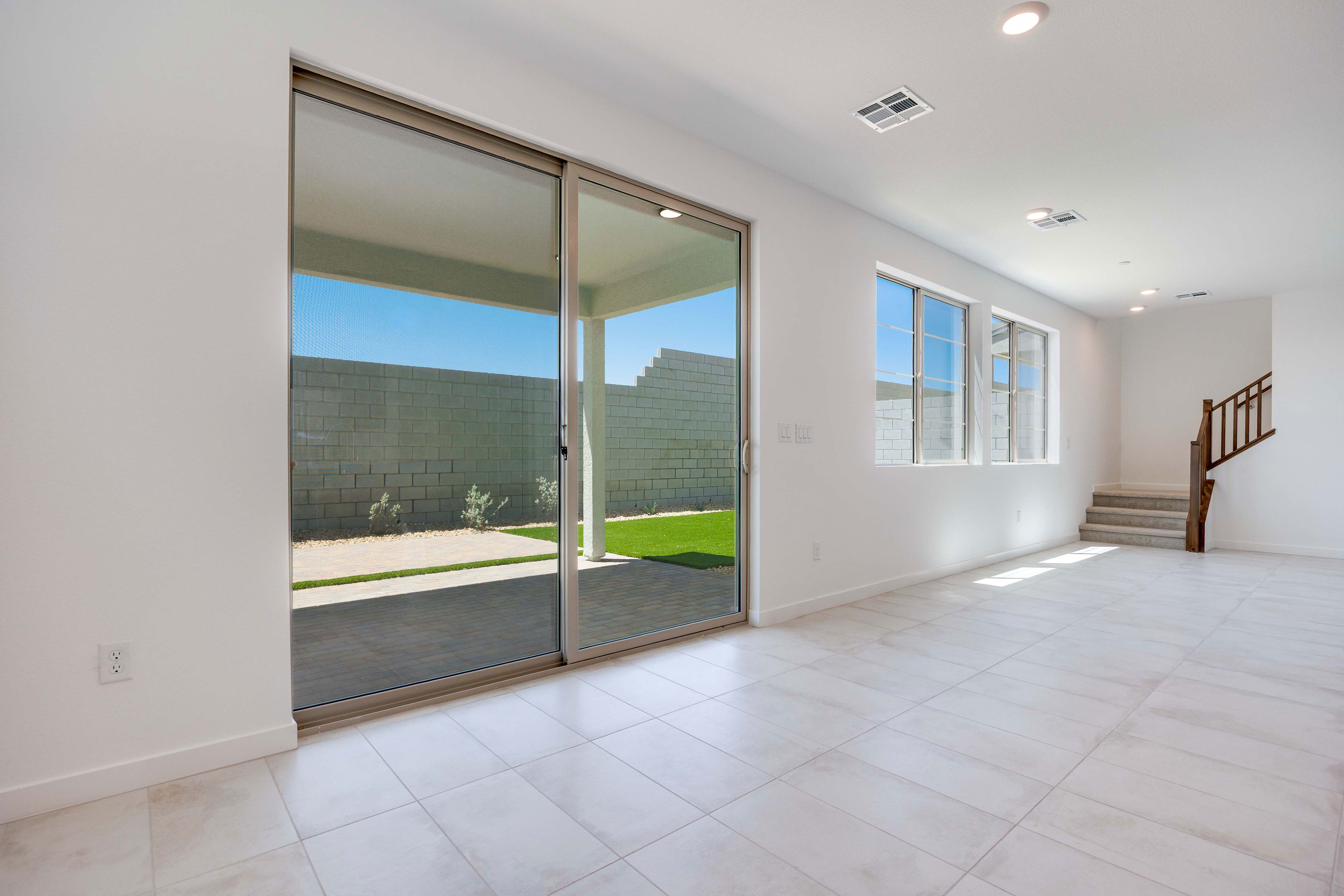 A bright, empty room with white tiled floors and a large sliding glass door in the foreground opens to a backyard with a patio and green grass, while the background features a staircase and windows.