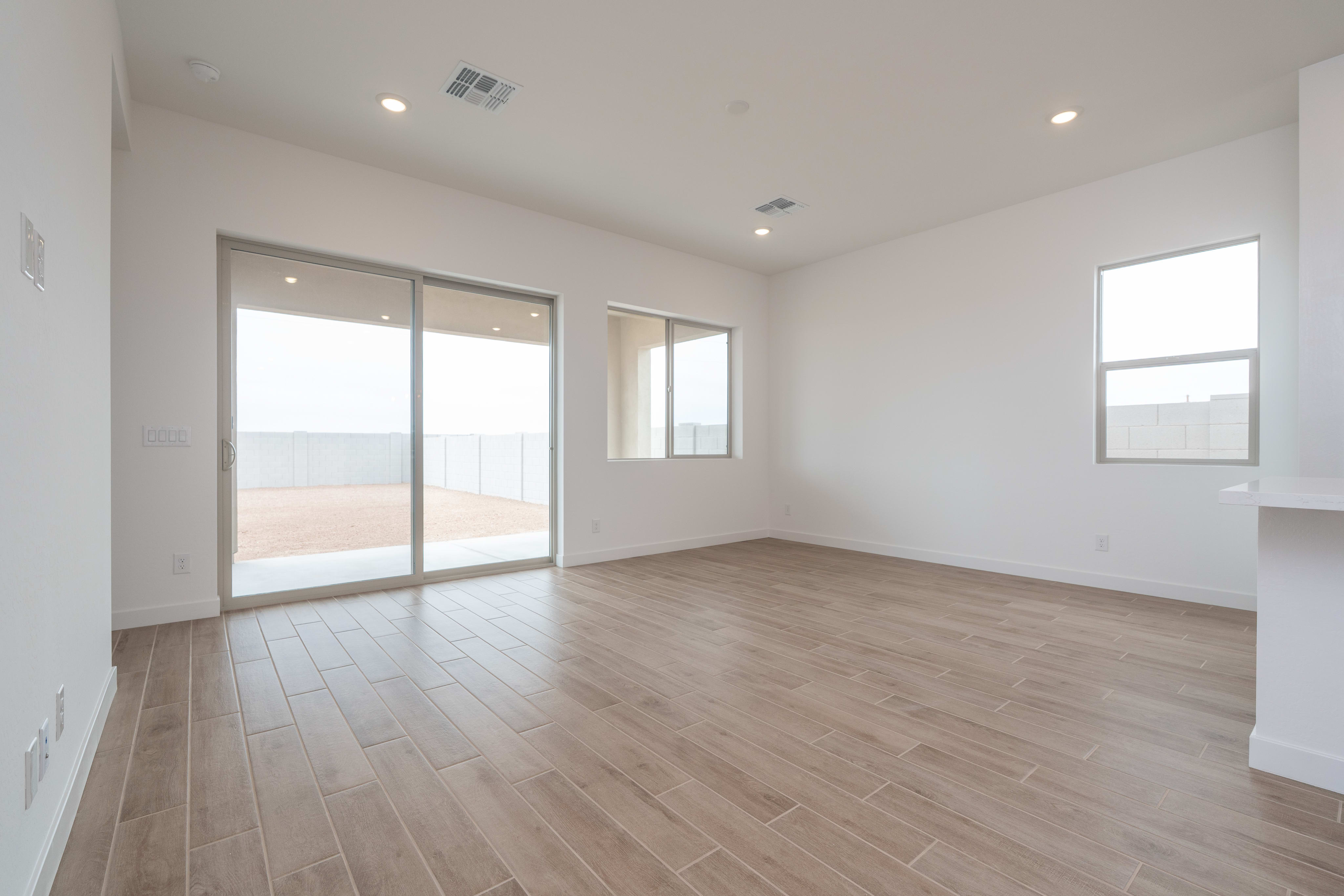 A spacious, empty room with light wood-look tile flooring in the foreground opens to a backyard through a large sliding glass door, with two windows and a bright, overcast sky visible in the background.