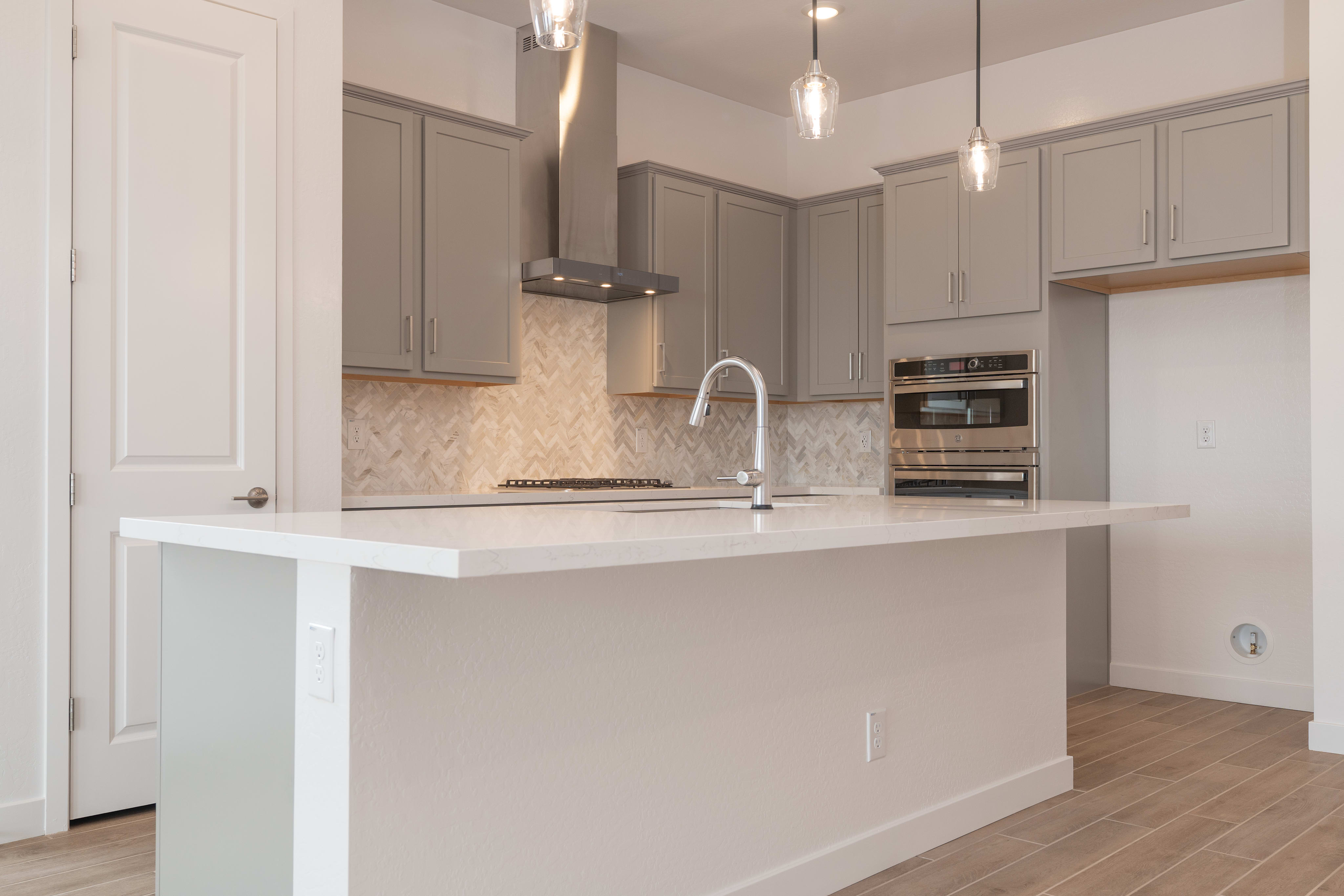 A modern kitchen island with a white countertop is in the foreground, while gray cabinets, stainless steel appliances, and a herringbone tile backsplash fill the background.