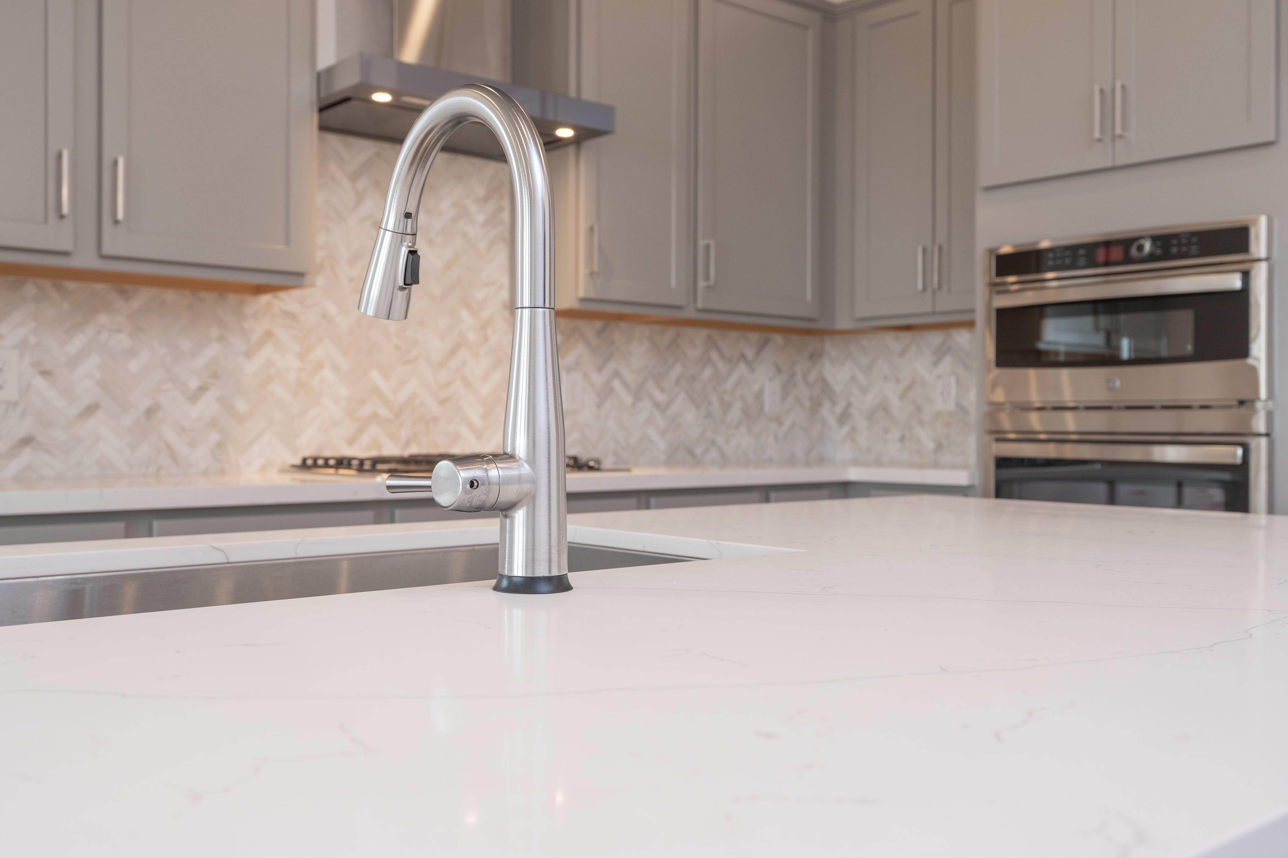 A modern kitchen features a sleek chrome faucet in the foreground and gray cabinets with stainless steel appliances in the background.