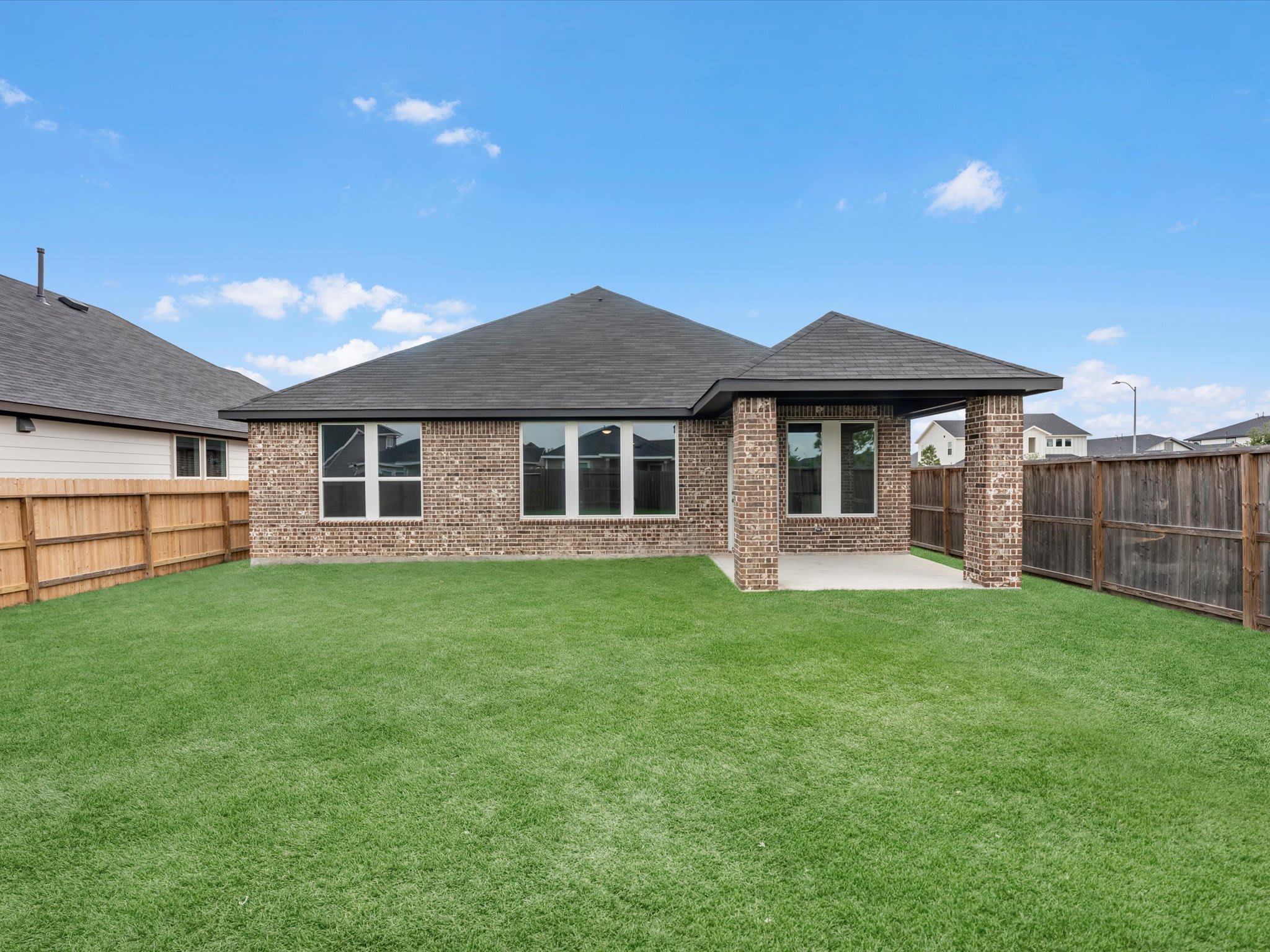 A freshly mowed green lawn stretches towards a brick house with a covered patio, all enclosed by a wooden fence under a bright blue sky with scattered clouds.