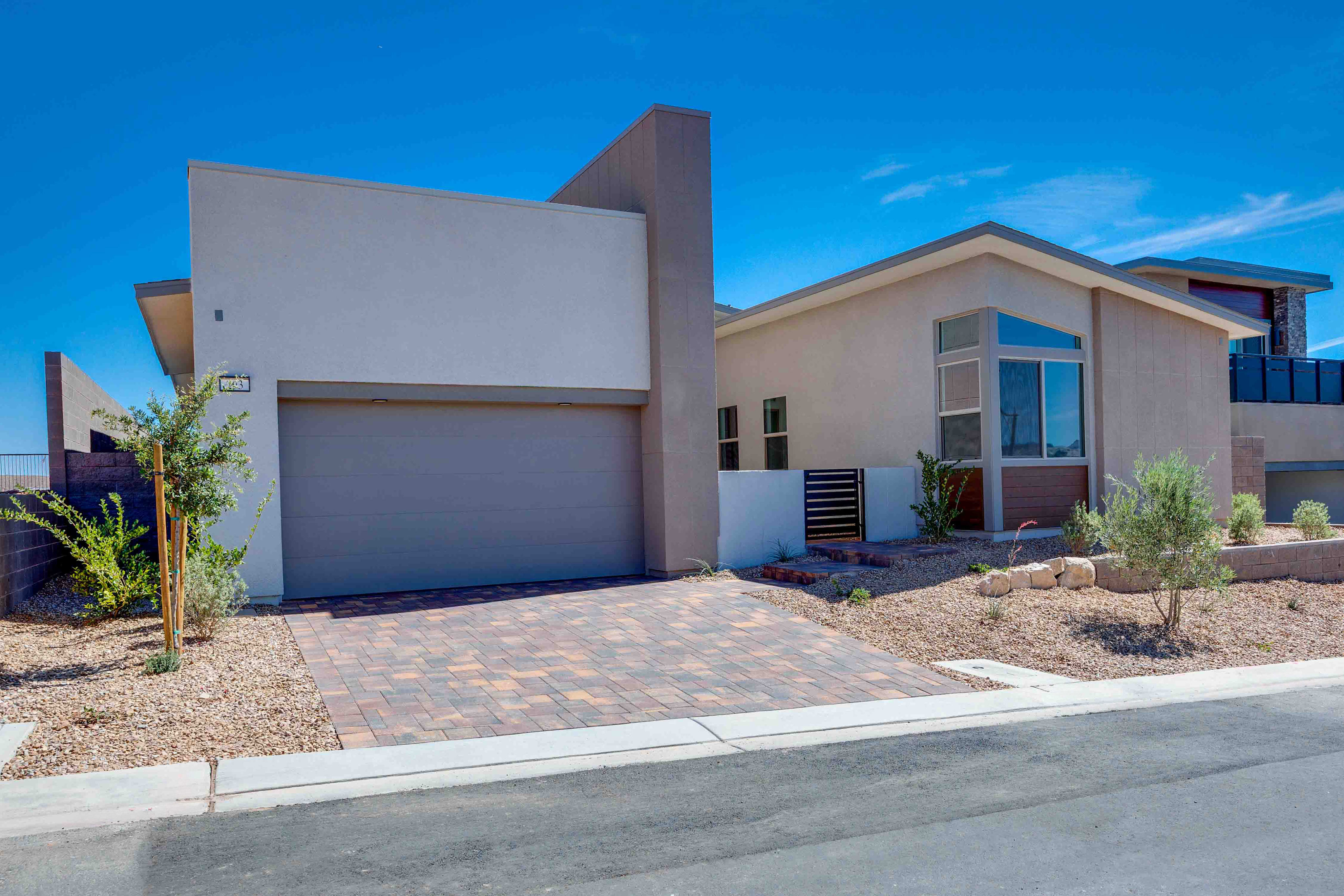 A modern beige house with a gray garage door sits on a paved driveway in the foreground, with a clear blue sky and distant hills in the background.