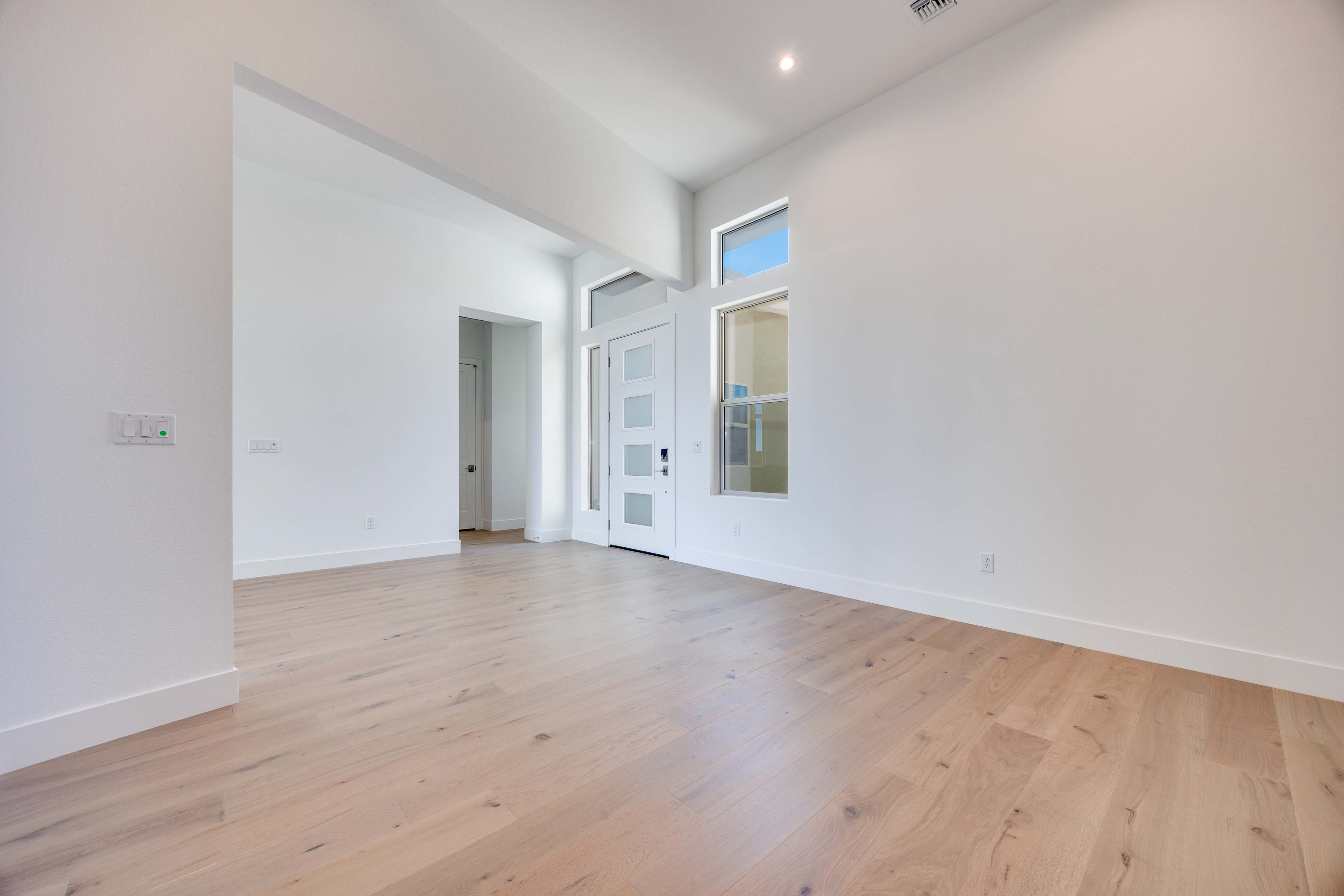 A bright, empty room with light wood floors in the foreground and a white door with glass panels and windows in the background.