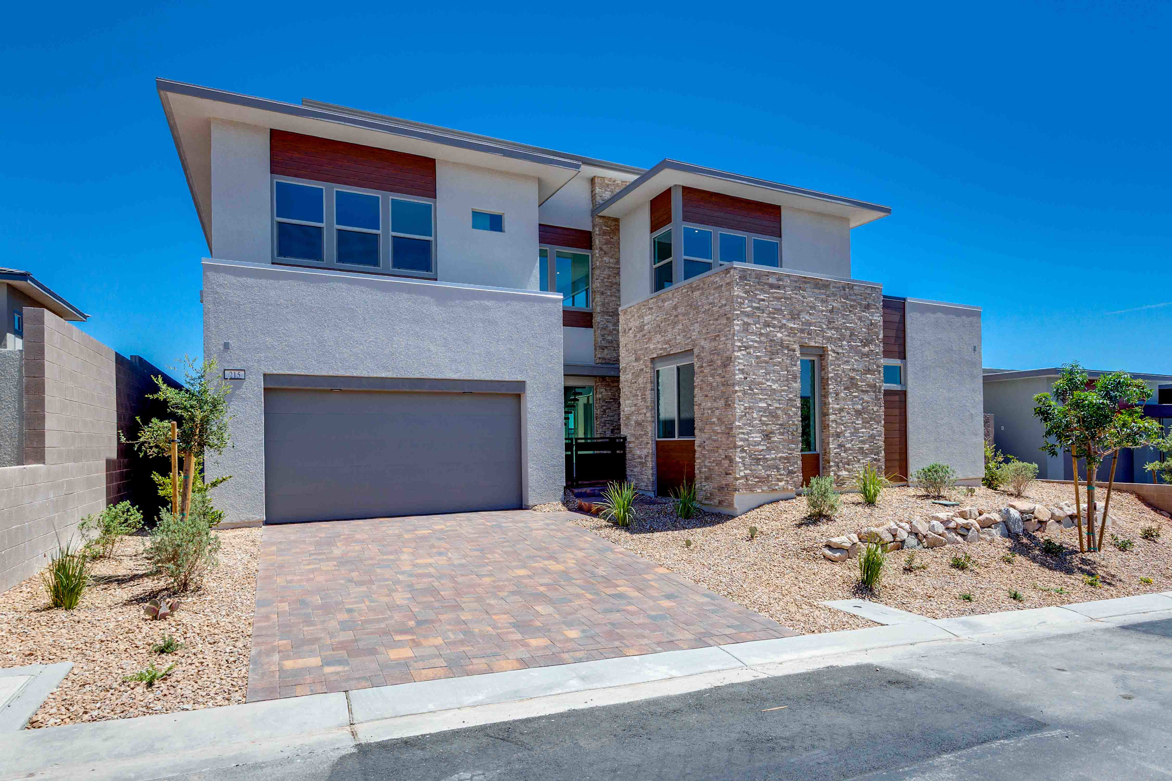A modern, two-story house with a stone accent wall and a paved driveway sits under a clear blue sky, with desert landscaping in the foreground and neighboring houses visible in the background.