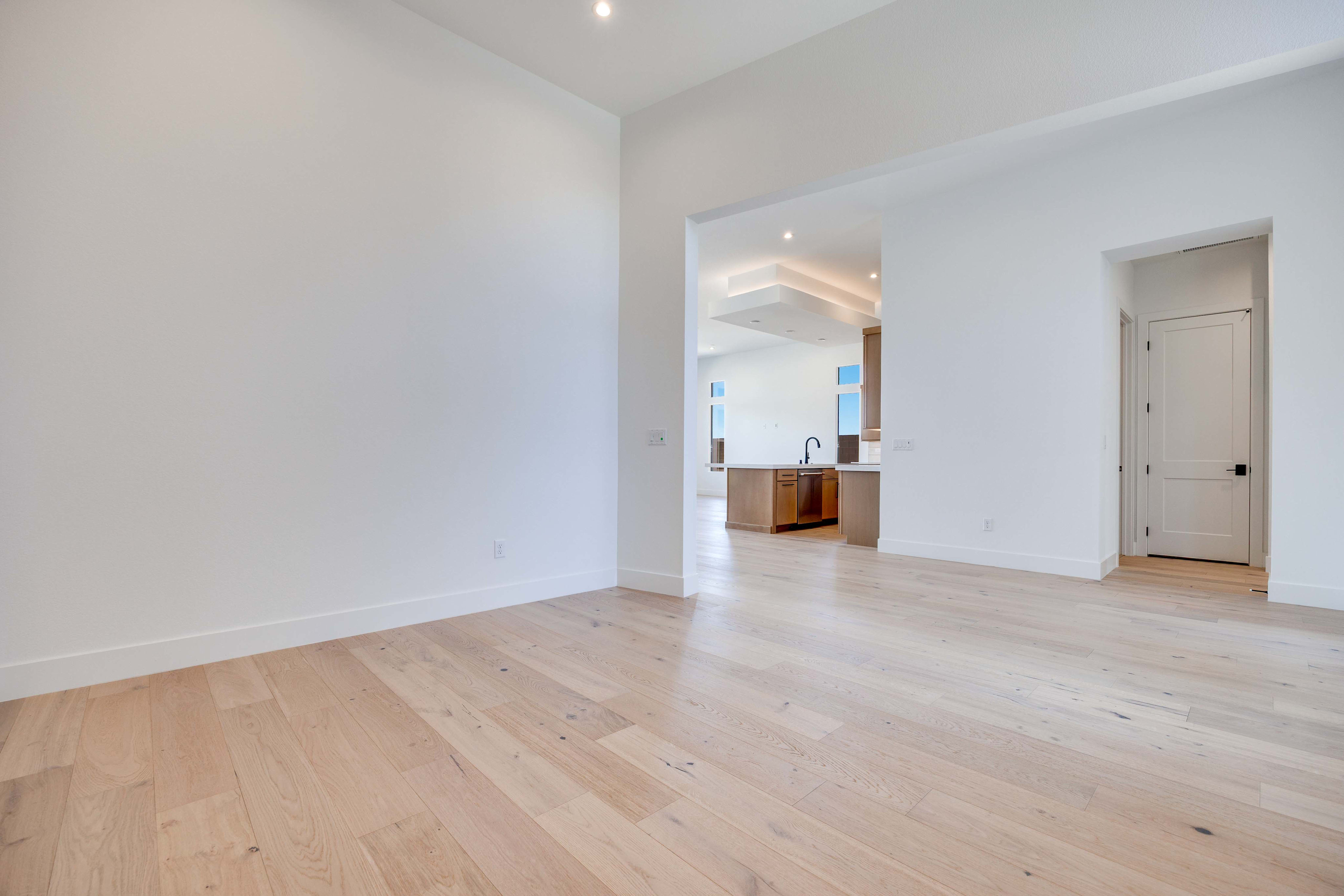 A bright, empty room with light wood floors in the foreground opens into a modern kitchen with recessed lighting in the background.