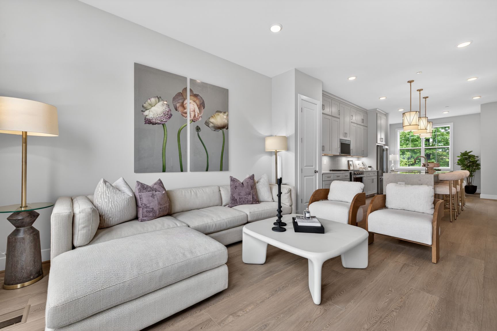 A modern living room with a sectional sofa and coffee table in the foreground, and a kitchen and dining area with a large window in the background.