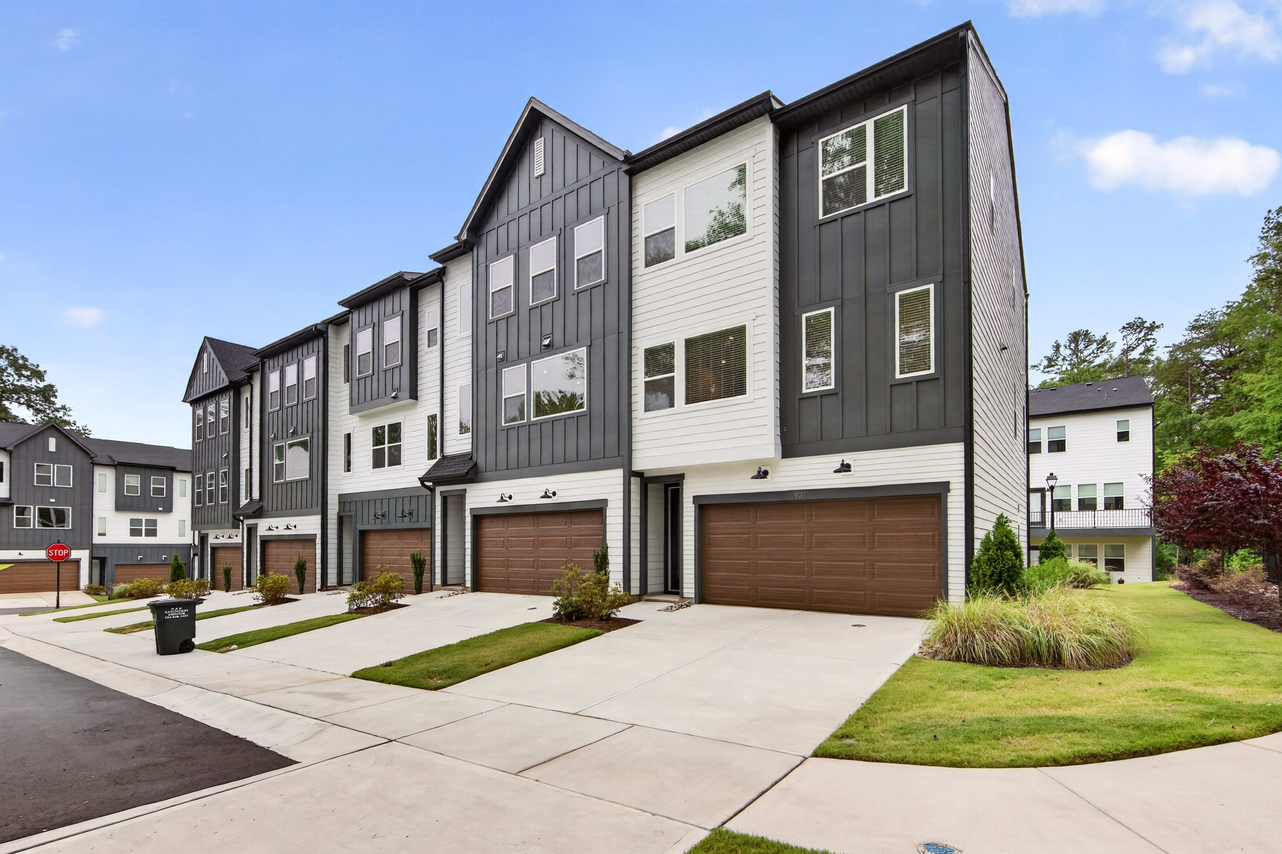 Modern townhouses with dark and white exteriors and brown garage doors line a paved street with manicured lawns in the foreground, while a backdrop of lush green trees and a bright blue sky completes the scene.