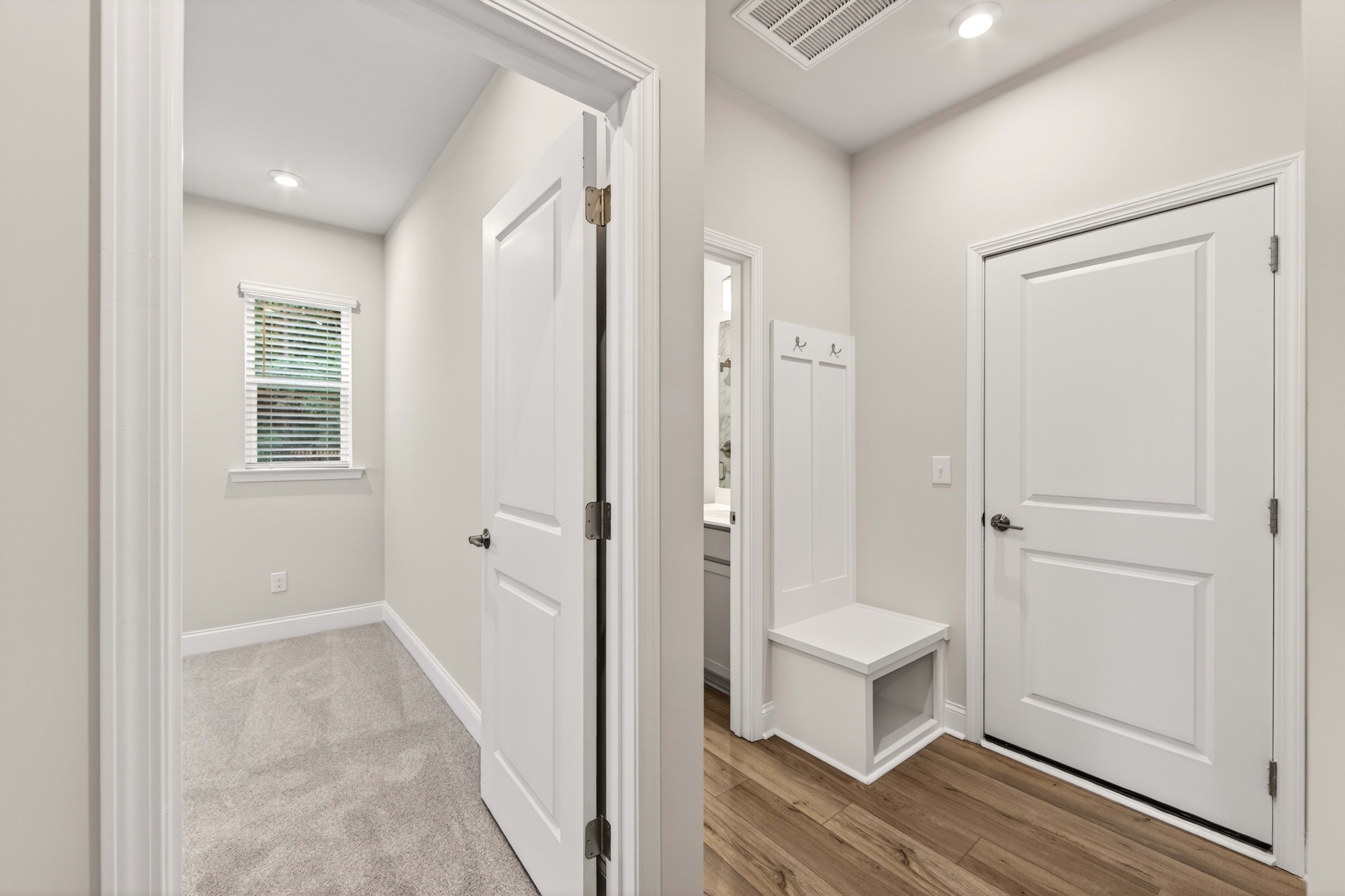 A bright hallway with a white bench and coat rack in the foreground leads to a carpeted room with a window in the background.