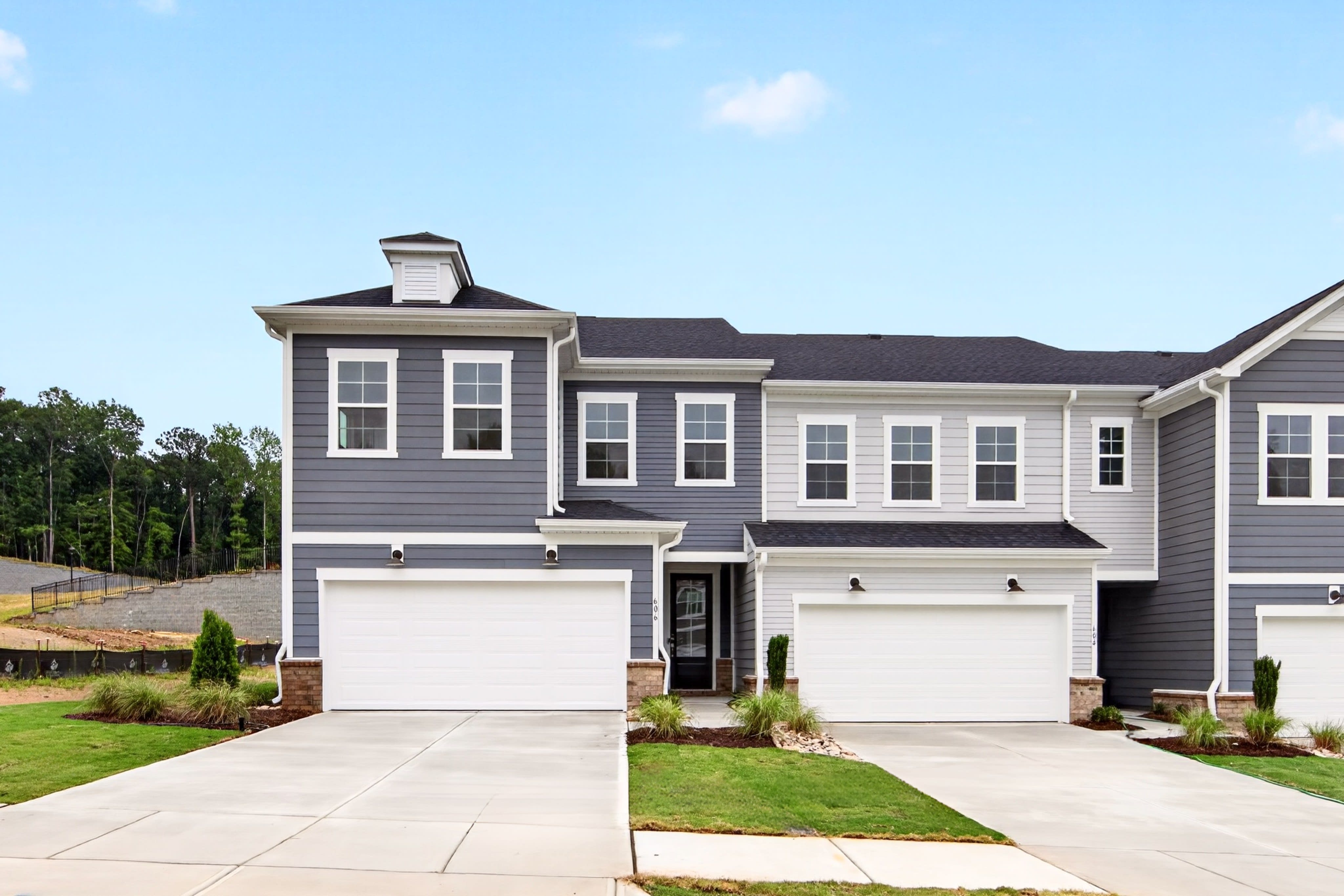 A row of modern townhouses with manicured lawns in the foreground stands against a backdrop of trees and a clear blue sky.