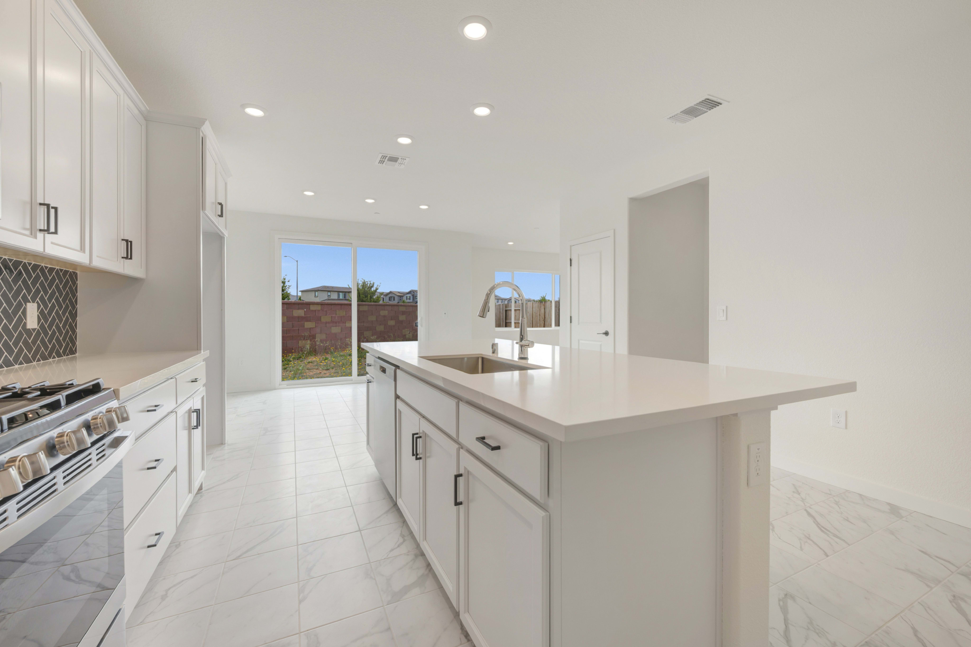 A modern white kitchen with a marble island in the foreground opens to a backyard with a clear blue sky in the background.