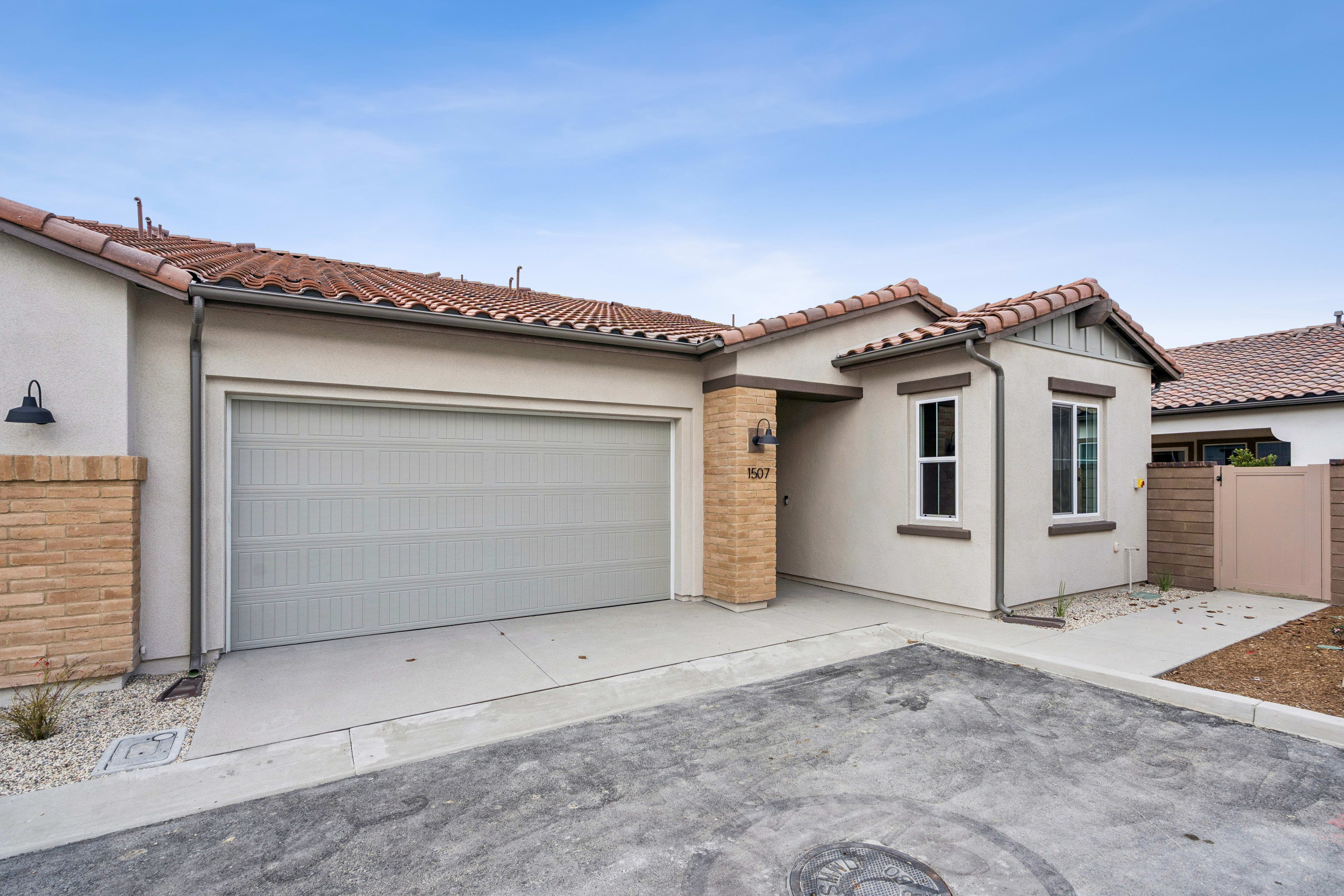 A modern beige house with a gray garage door and a tiled roof sits under a cloudy blue sky, with a paved driveway in the foreground.
