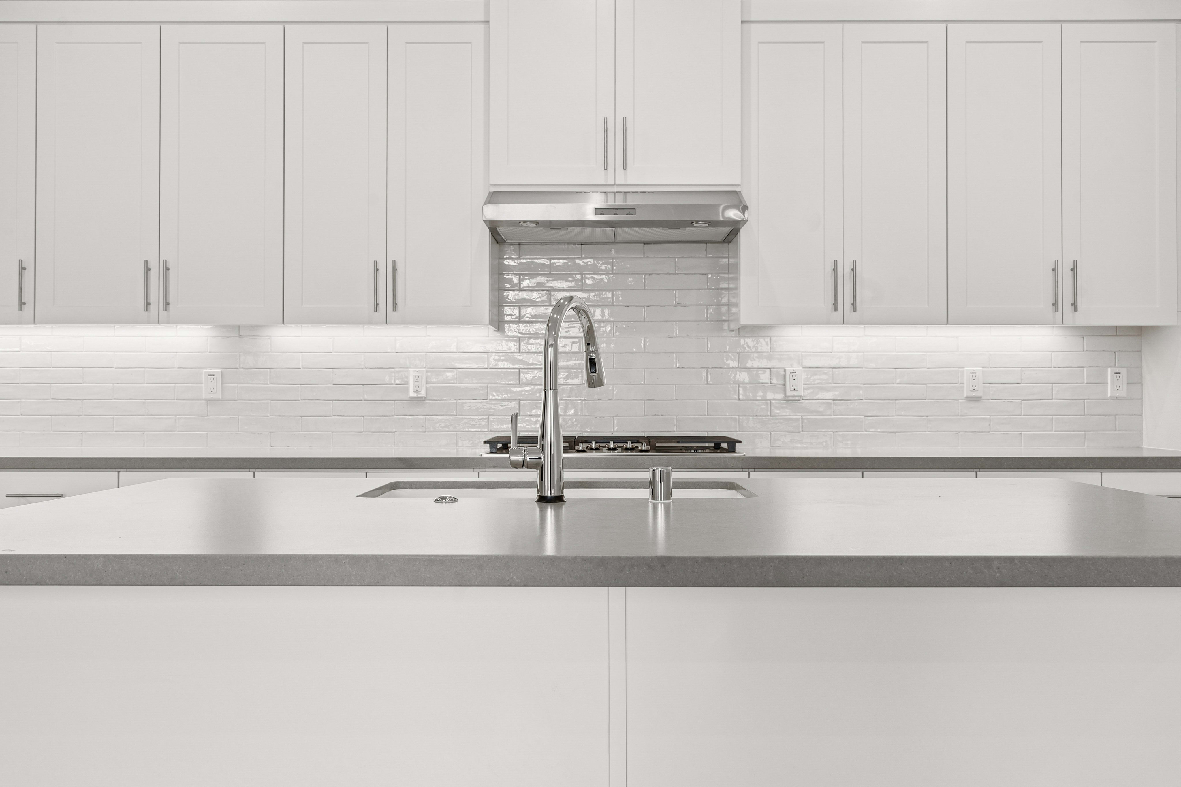 A modern kitchen island with a sleek countertop and faucet is in the foreground, while white cabinets and a tiled backsplash fill the background.