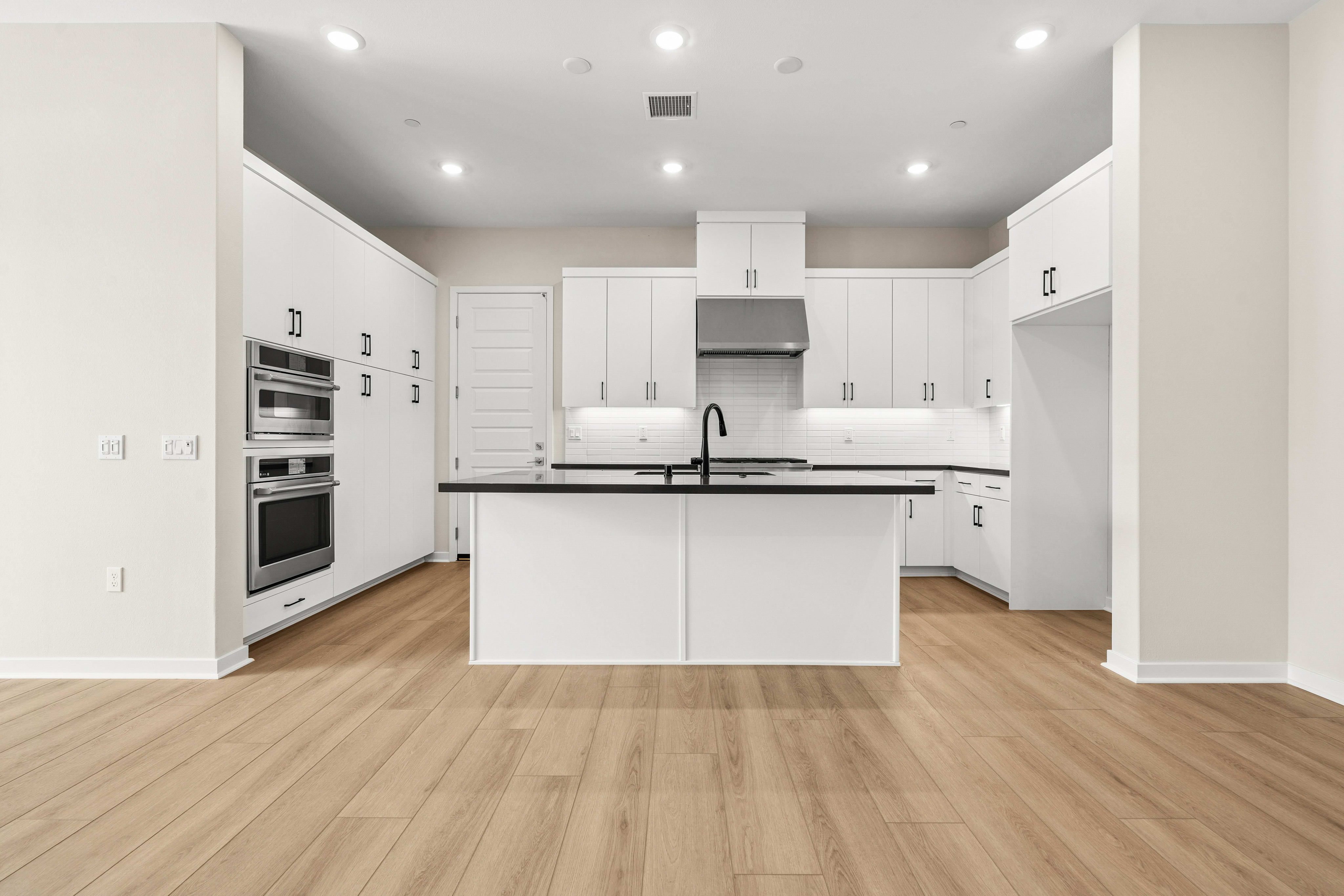 A modern white kitchen with a central island and stainless steel appliances is visible in the background, while the foreground features a light wood floor.