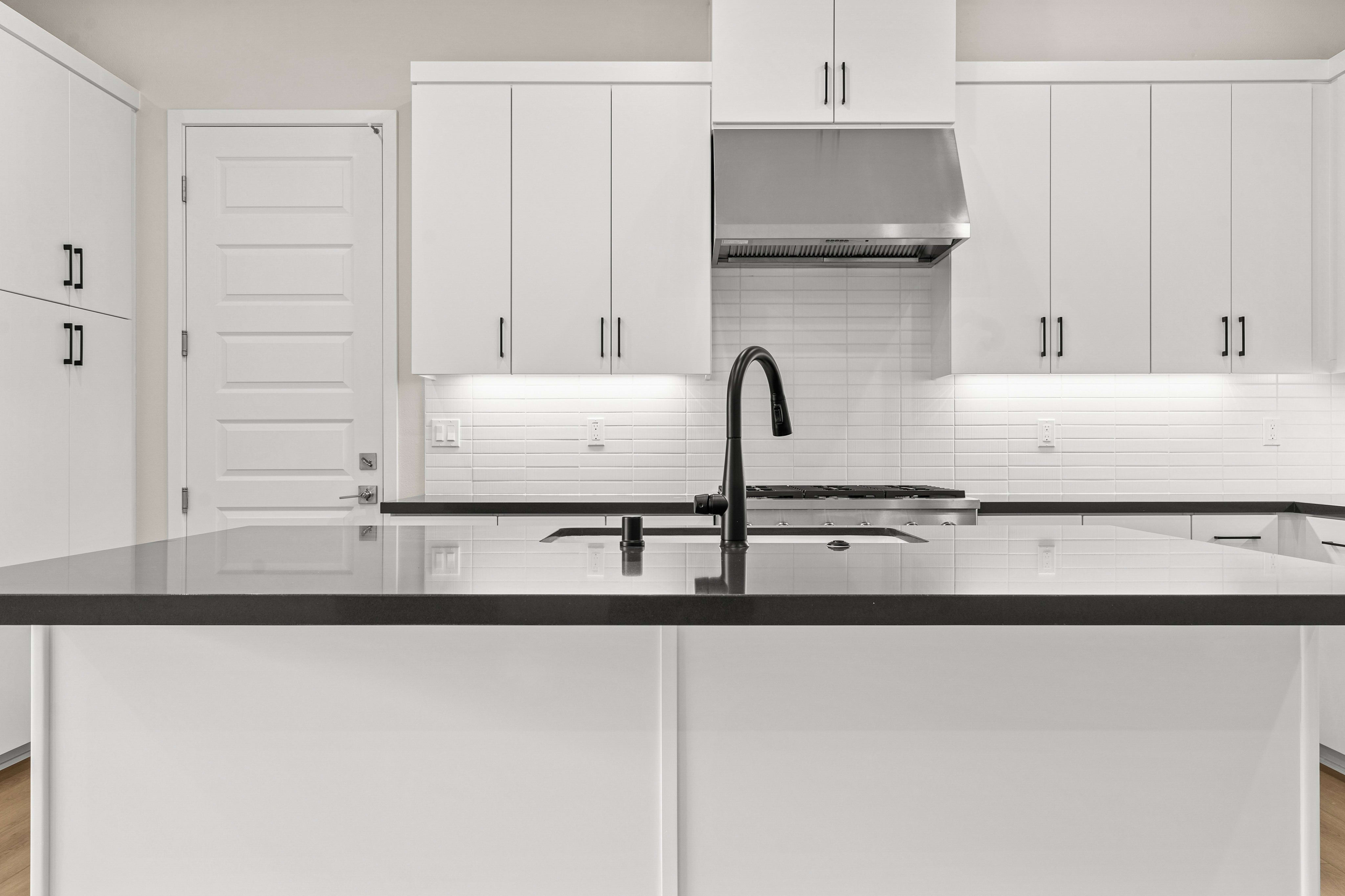 A sleek black countertop island with a modern faucet sits in the foreground of a bright white kitchen with stainless steel appliances and a white subway tile backsplash.