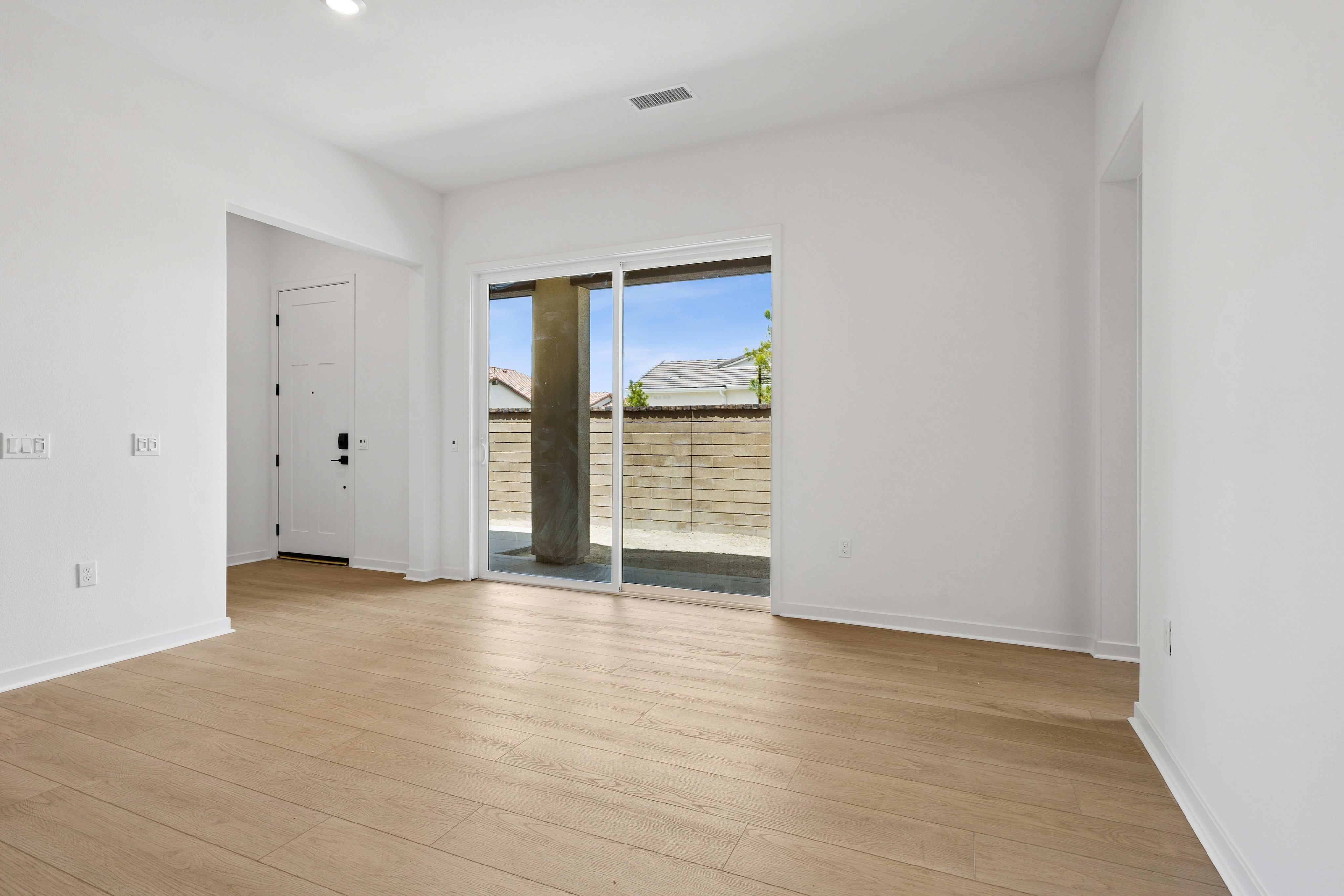 A bright, empty room with light wood flooring in the foreground opens to a patio with a concrete pillar and a view of a fence and sky in the background.