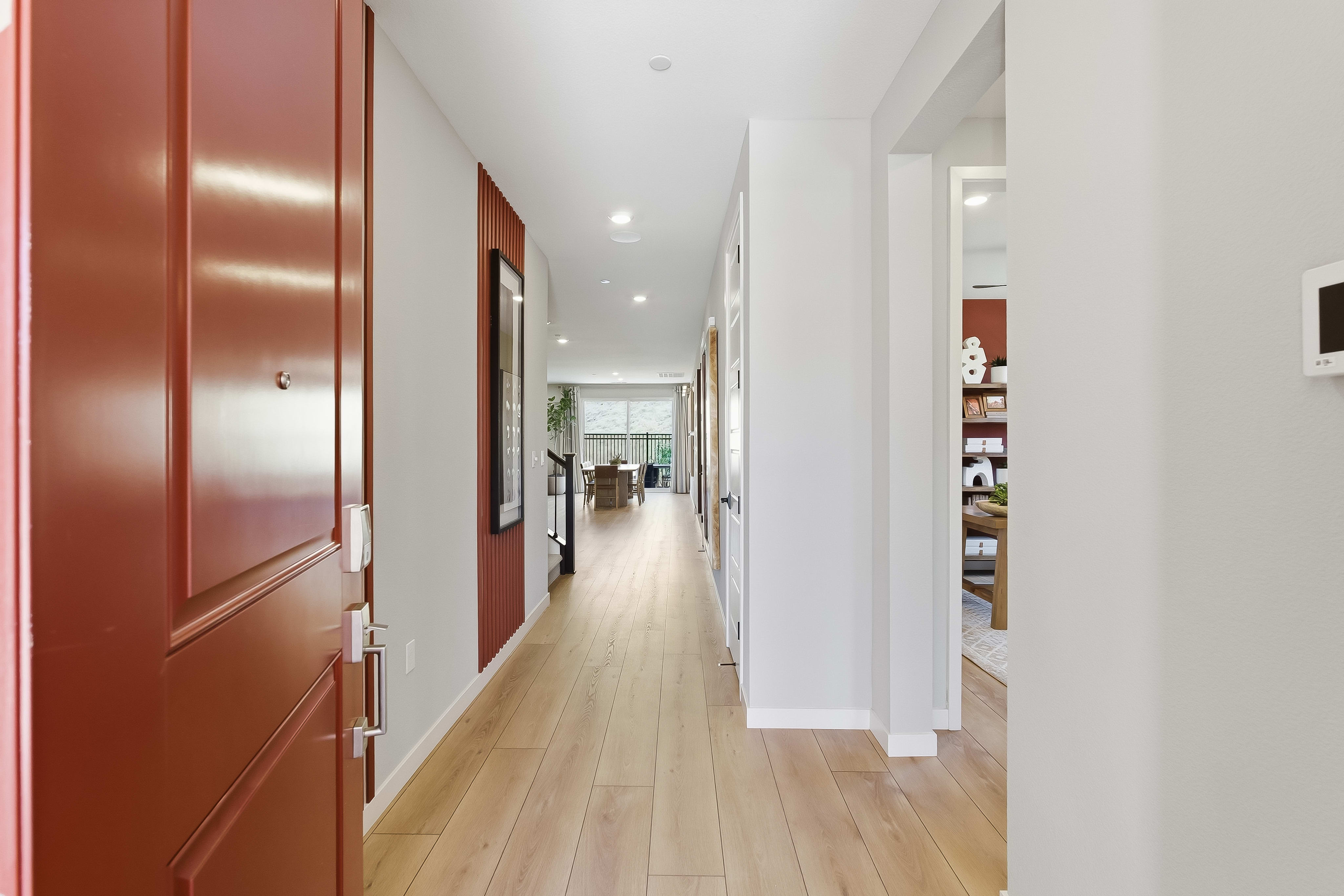 A red front door opens into a bright hallway with light wood floors, leading to a living area with large windows in the background.