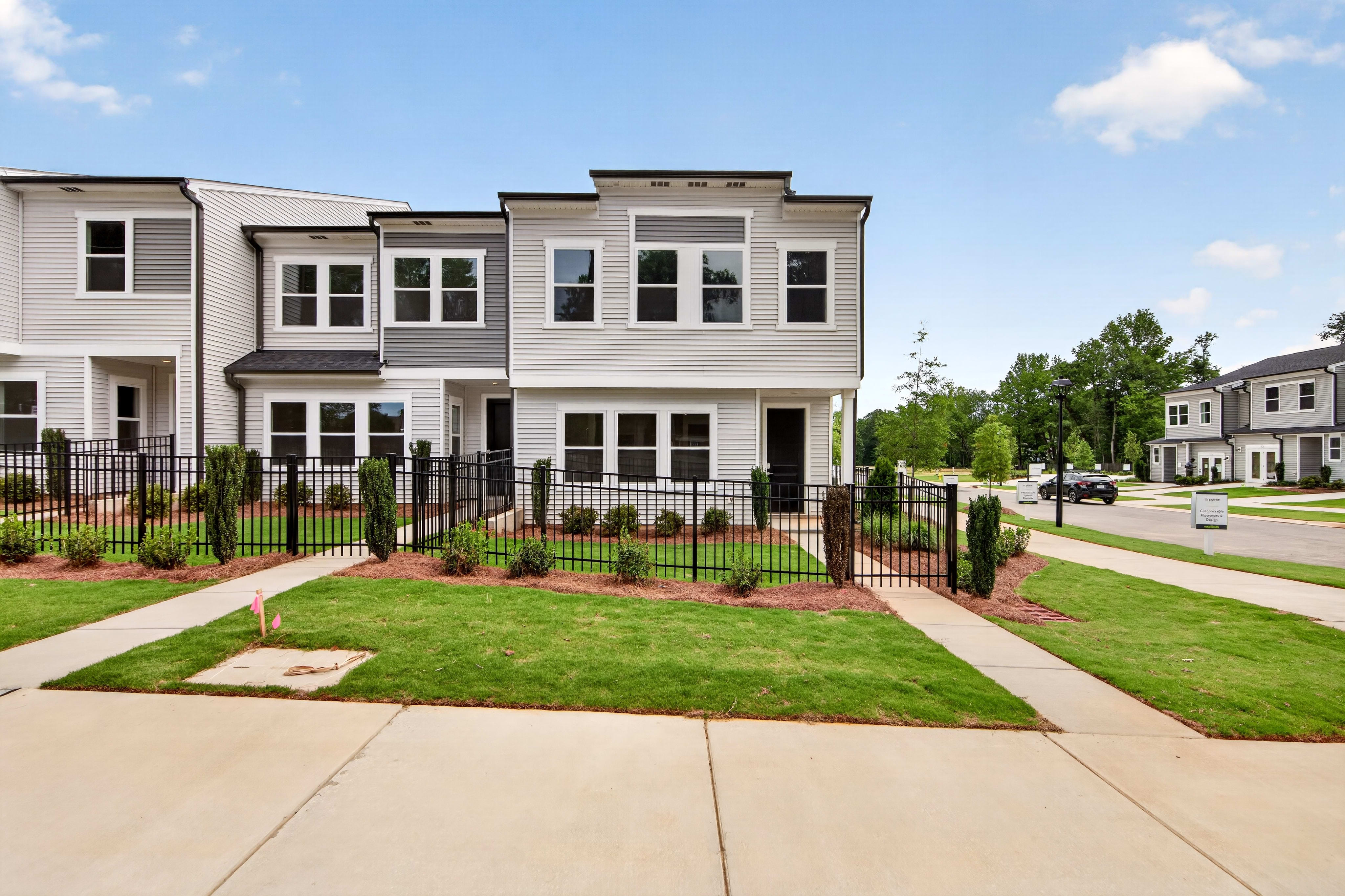 A row of modern townhouses with manicured lawns and black metal fences in the foreground stands against a backdrop of trees and a clear blue sky.